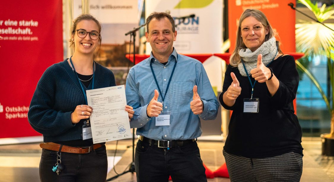 KI generiert: Das Bild zeigt drei Personen, die Daumen hoch zeigen, eine hält ein Dokument in der Hand. Sie wirken glücklich und befinden sich in einem Innenraum mit Informations- und Werbeschildern im Hintergrund.