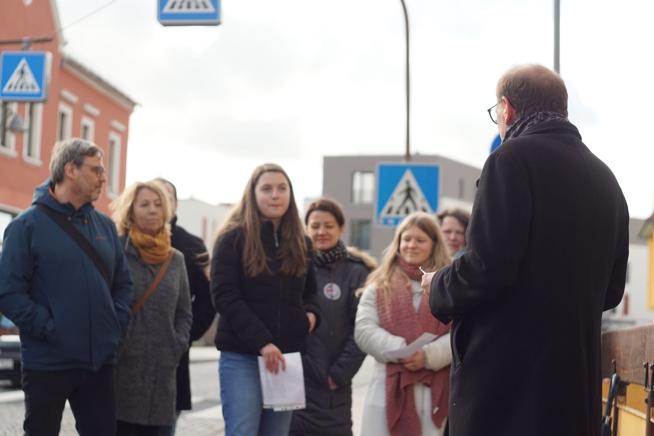 KI generiert: Eine Gruppe von Menschen steht im Freien, während eine Person im Vordergrund ihnen gegenüberzutreten scheint, möglicherweise sprechend oder erklärend. Im Hintergrund sind Verkehrszeichen für einen Fußgängerüberweg sichtbar.