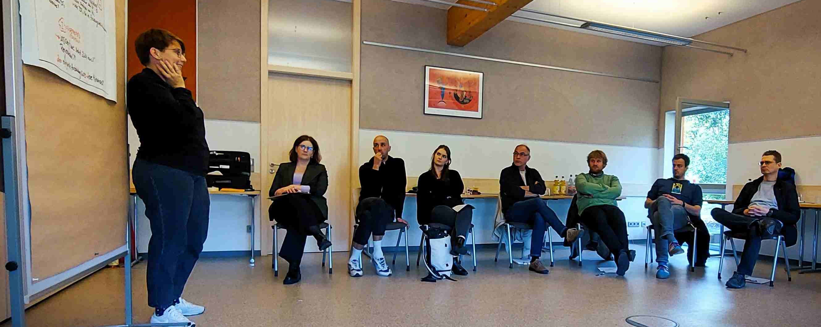 KI generiert: Das Bild zeigt eine Gruppe von Menschen, die in einem Stuhlkreis in einem Raum sitzen, während eine Person vorne steht und möglicherweise eine Präsentation hält oder etwas erklärt. Der Raum hat eine entspannte, informelle Atmosphäre.