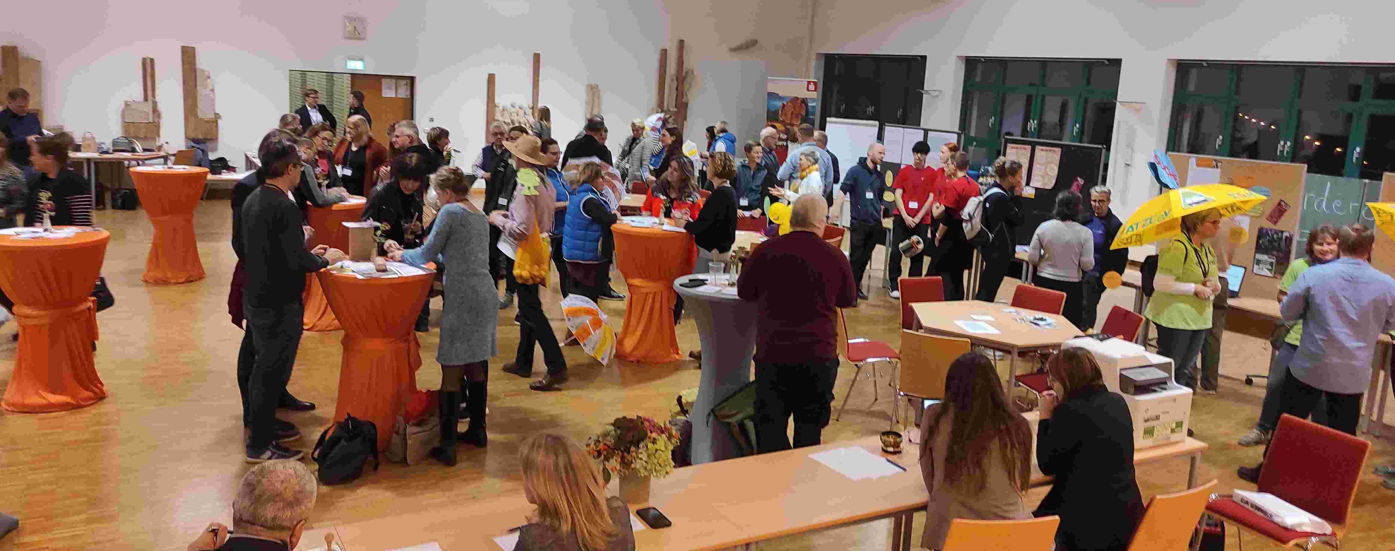 KI generiert: Das Bild zeigt eine Veranstaltung mit Menschen, die in einem Raum an Stehtischen stehen oder sich unterhalten. Im Hintergrund sind Informationsstände zu sehen.