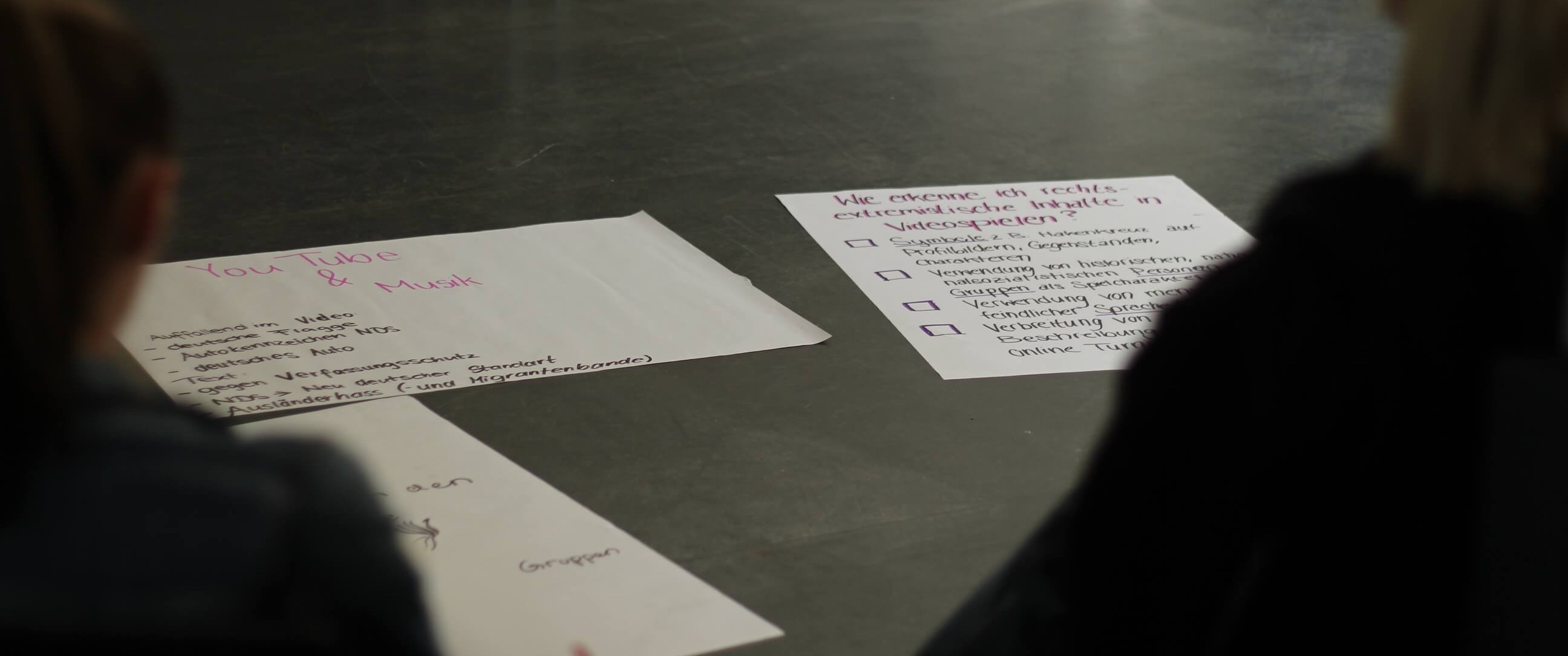 KI generiert: Das Bild zeigt zwei große Papierbögen auf dem Boden, die mit handschriftlichen Notizen versehen sind. Personen im Vordergrund schauen auf die Bögen, die scheinbar Themen wie "YouTube & Musik" und Entscheidungsfindung behandeln.