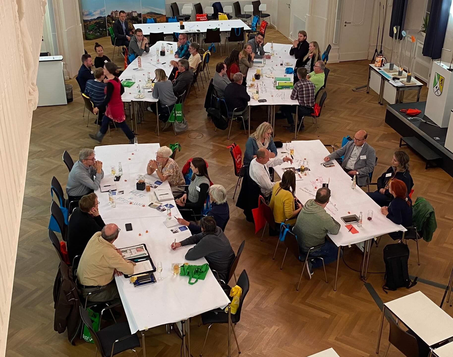 KI generiert: Das Bild zeigt eine Versammlung von Menschen, die an mehreren Tischen in einem großen Raum sitzen und diskutieren oder arbeiten. Der Raum ist mit Holzboden ausgelegt und hat eine offene, formelle Atmosphäre.