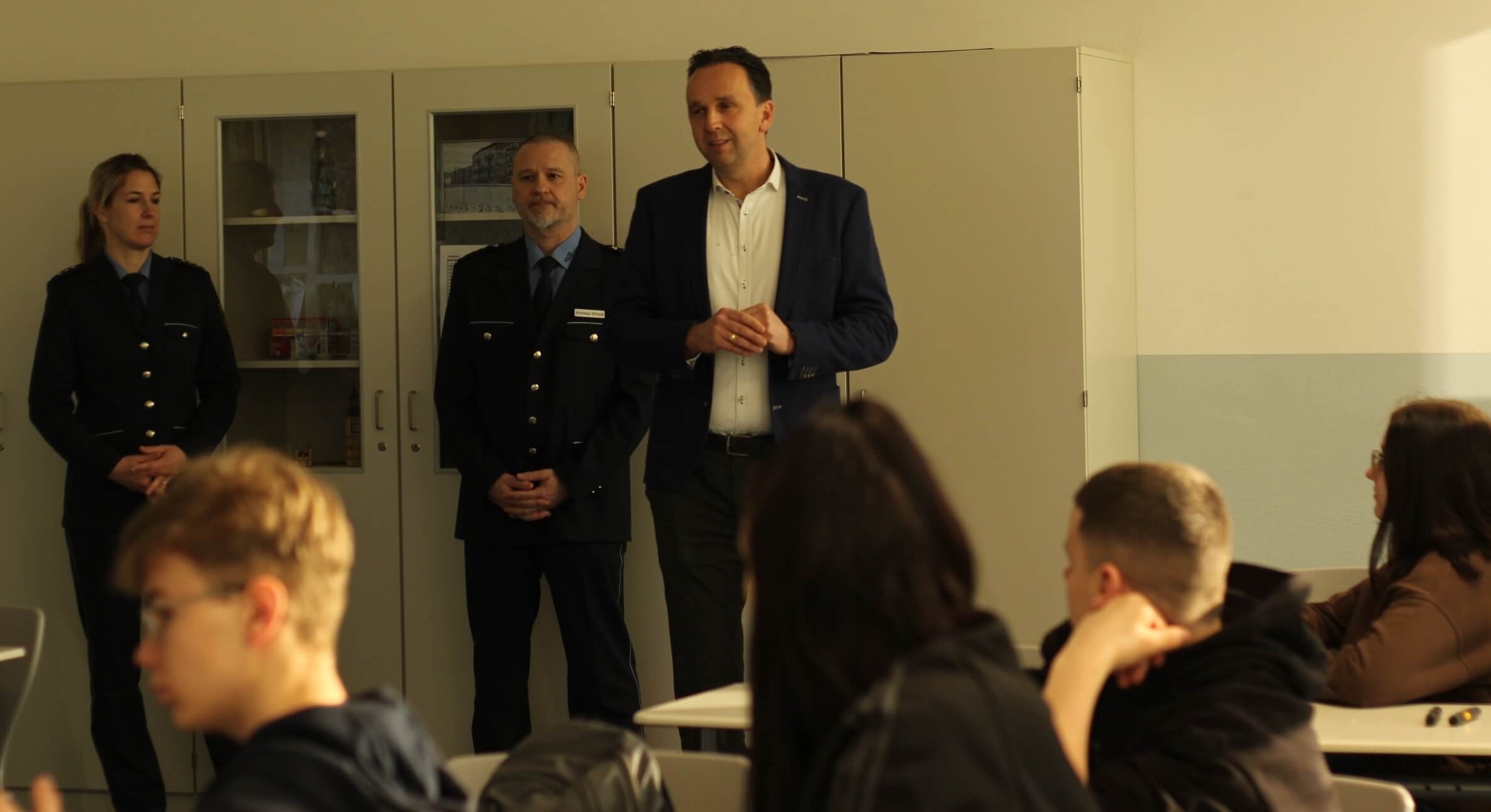 KI generiert: Das Bild zeigt drei Personen, darunter zwei in Uniform, die vor einer Gruppe von sitzenden Menschen in einem Klassenzimmer stehen. Es scheint eine Präsentation oder ein Vortrag stattzufinden.