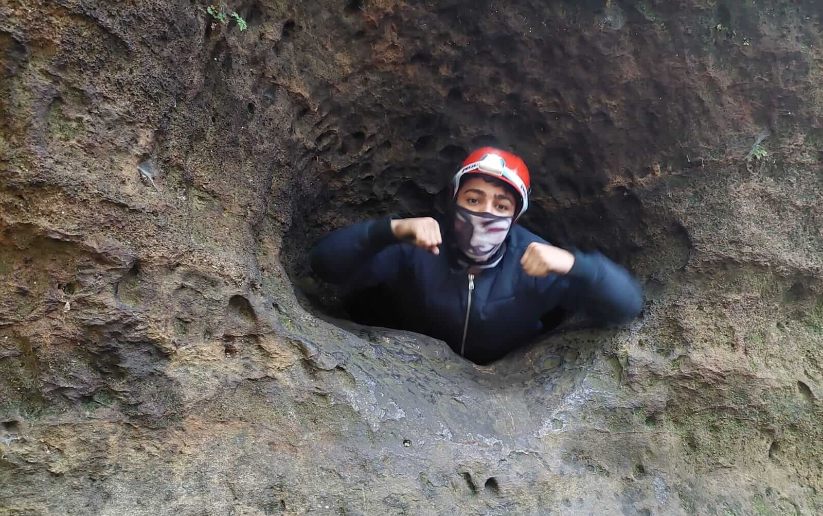 KI generiert: Das Bild zeigt eine Person, die aus einer größeren Öffnung in einer Felswand herausblickt, mit einem roten Helm und einem Gesichtstuch. Die Person hat die Arme angewinkelt, als ob sie eine kraftvolle Geste macht.