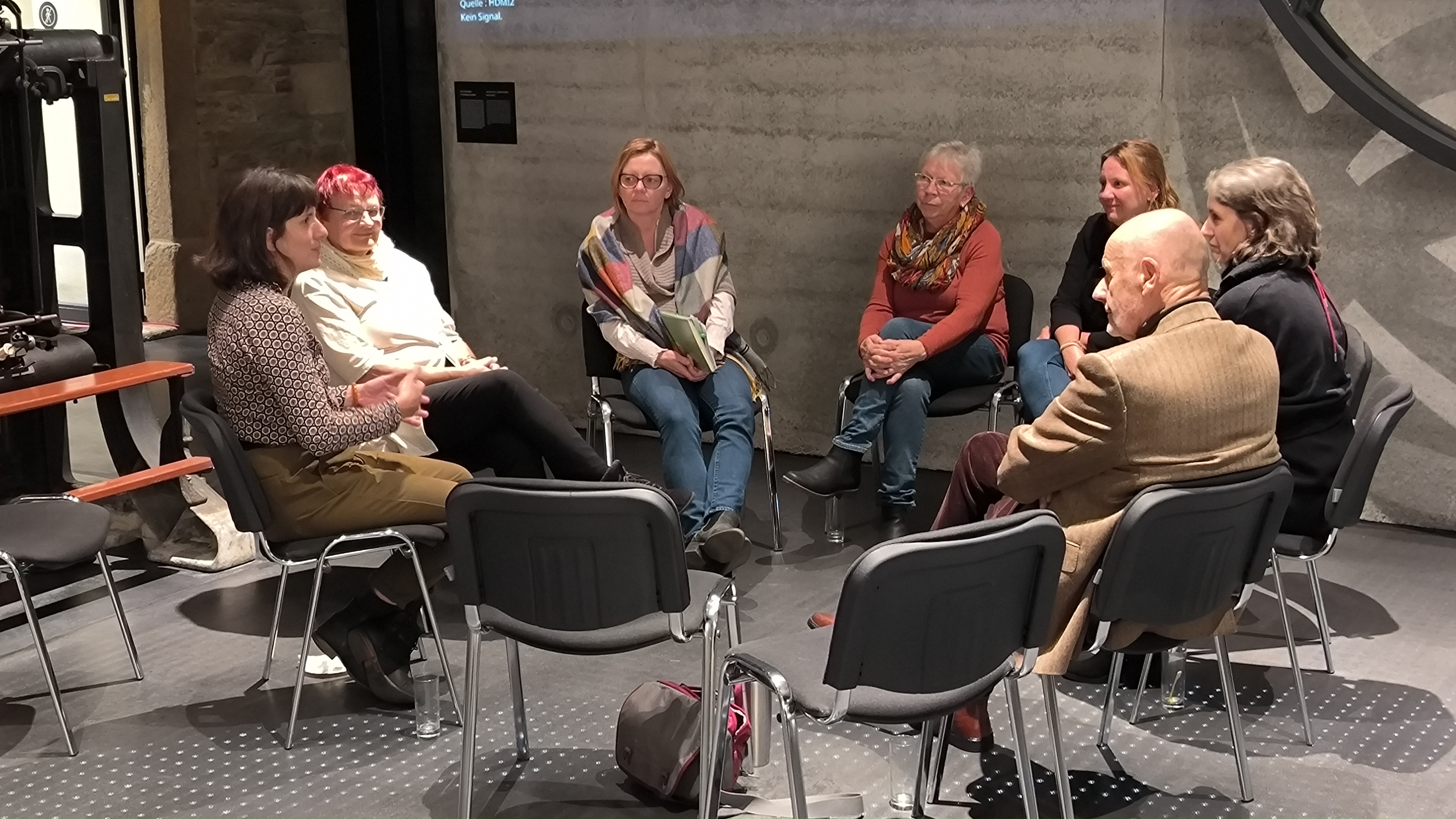 KI generiert: Eine Gruppe von sieben Personen sitzt in einem Stuhlkreis in einem Raum und unterhält sich. Die Atmosphäre wirkt entspannt und der Raum hat eine moderne Einrichtung mit Betonwänden.