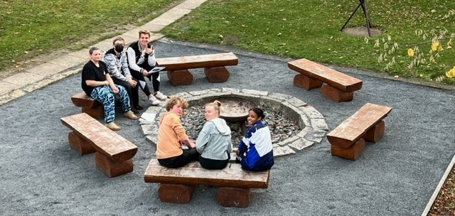 KI generiert: Eine Gruppe von Menschen sitzt draußen auf Holzbänken um eine Feuerstelle herum. Die Stimmung wirkt freundlich und entspannt.