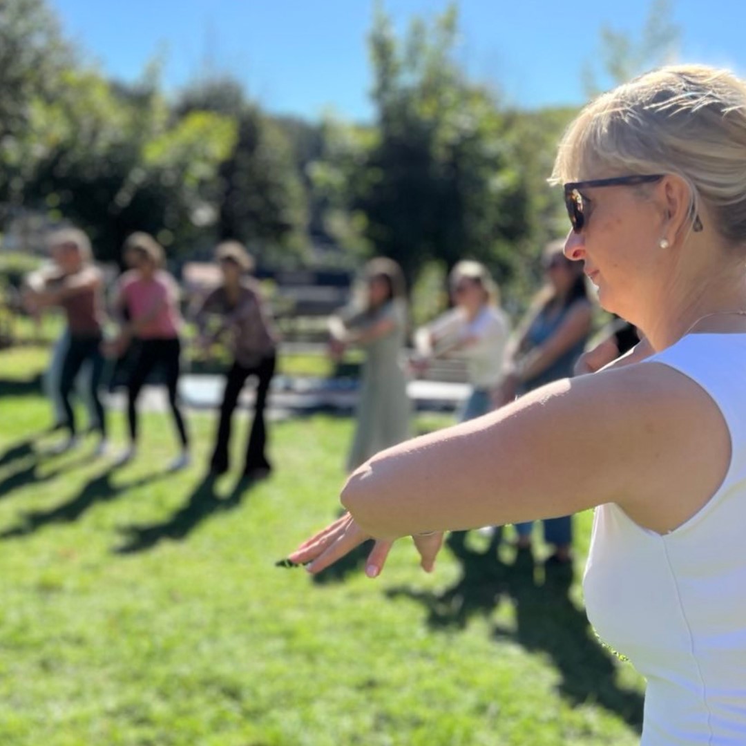 KI generiert: Das Bild zeigt eine Frau im Vordergrund, die in einem grünen Park steht und einen entspannten Bewegungsablauf ausführt. Im Hintergrund sind weitere Personen zu sehen, die offenbar an einer ähnlichen Aktivität teilnehmen.