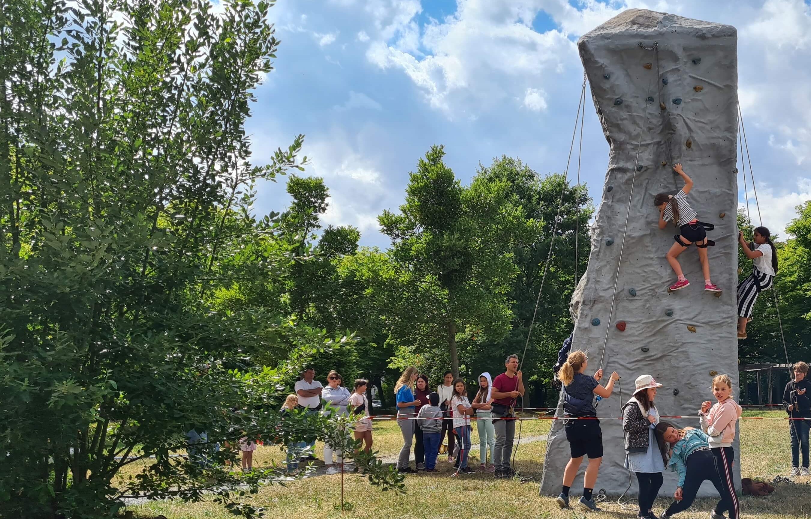 KI generiert: Das Bild zeigt eine Gruppe von Menschen im Freien, die einer Kletterwand zusehen, während einige Personen darauf klettern. Die Szene spielt sich in einer parkähnlichen Umgebung mit Bäumen im Hintergrund ab.