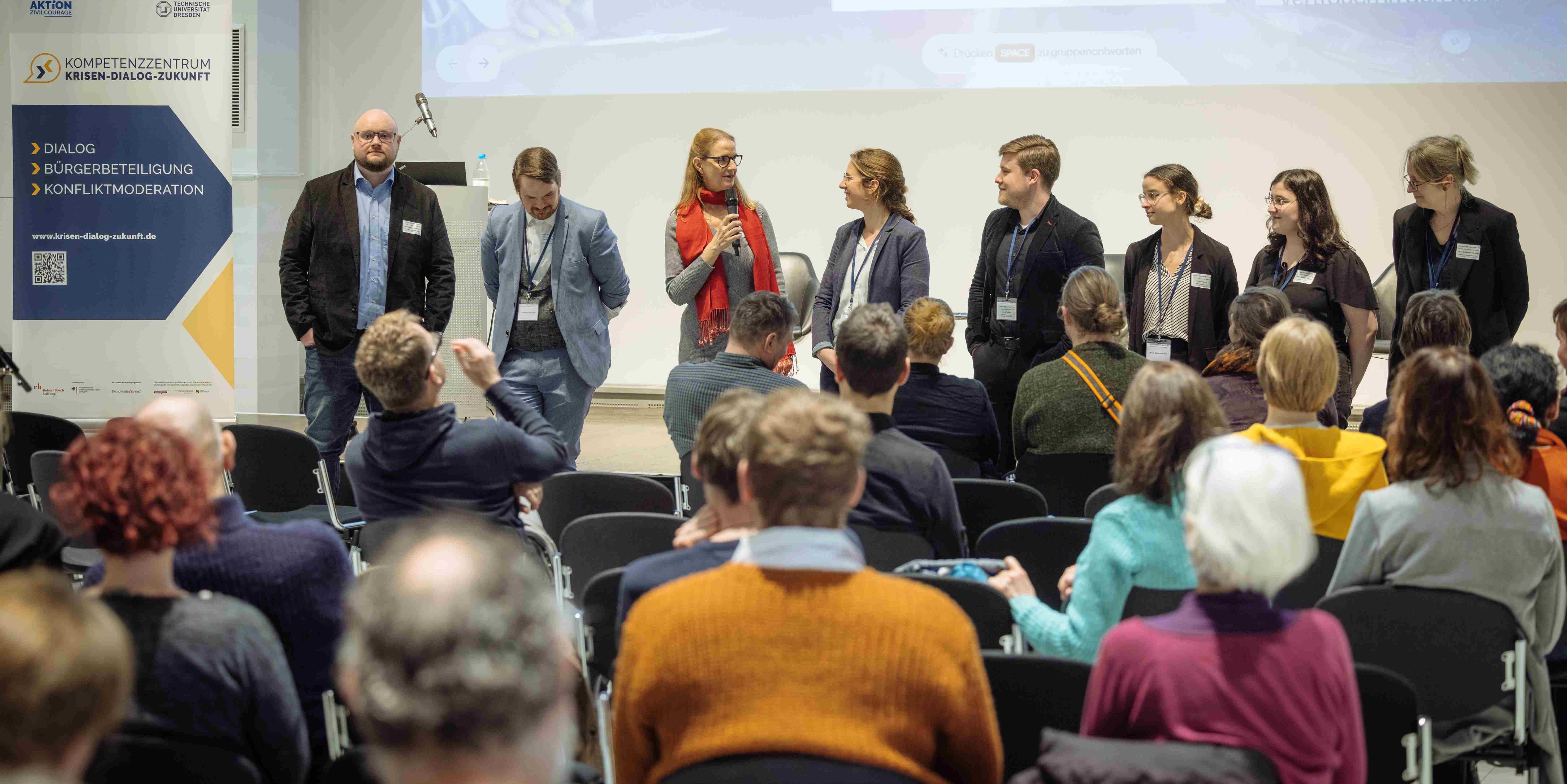 KI generiert: Das Bild zeigt eine Gruppe von Menschen, die sich vorne in einem Veranstaltungsraum vor einer sitzenden Menge befinden, während eine Person ein Mikrofon in der Hand hält und spricht. Im Hintergrund ist ein Banner des "Kompetenzzentrums Krisen-Dialog-Zukunft" sichtbar.