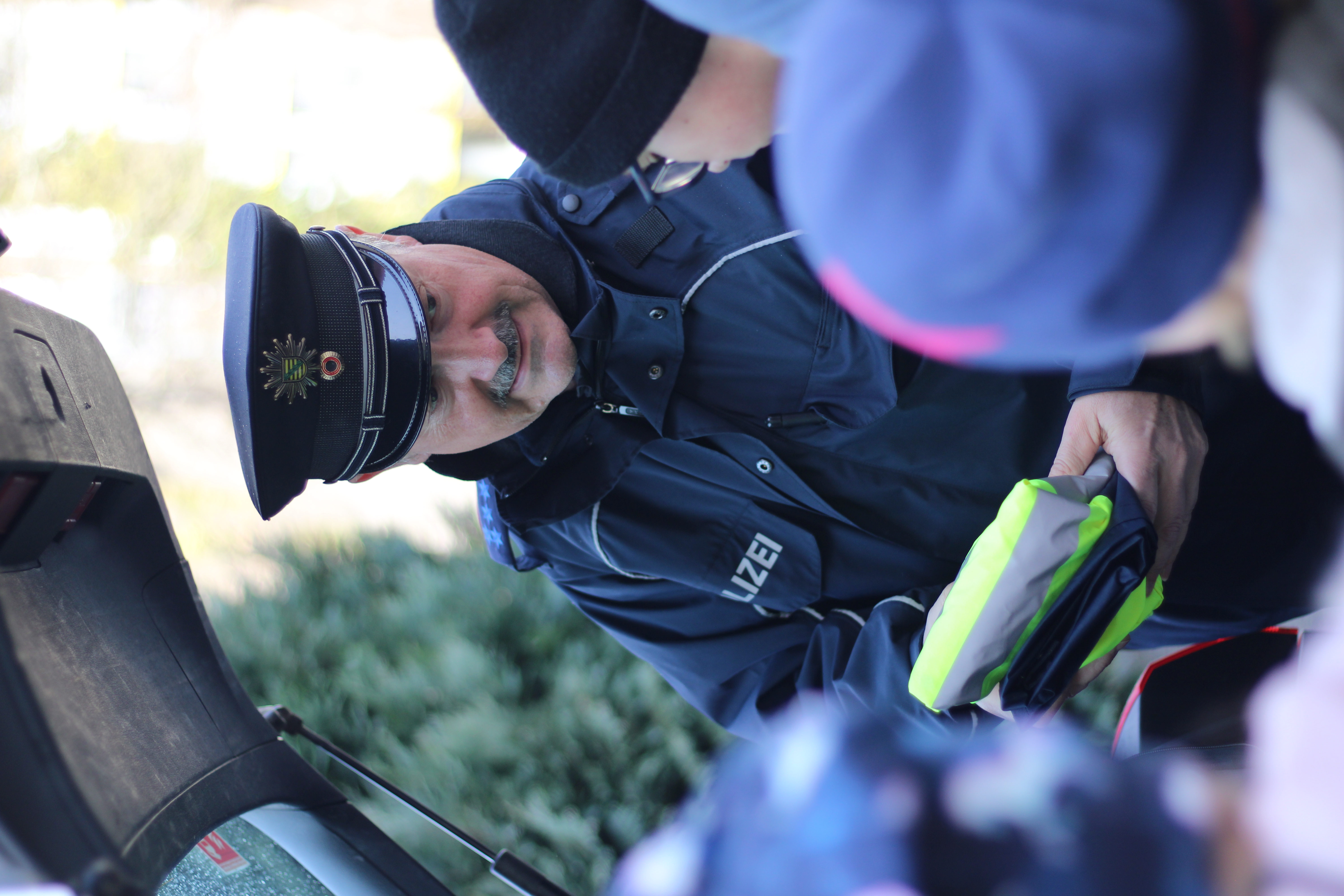 KI generiert: Ein Polizist interagiert mit einer Gruppe von Menschen und hält dabei ein reflektierendes Objekt in den Händen. Im Hintergrund ist ein geöffneter Kofferraum zu sehen.