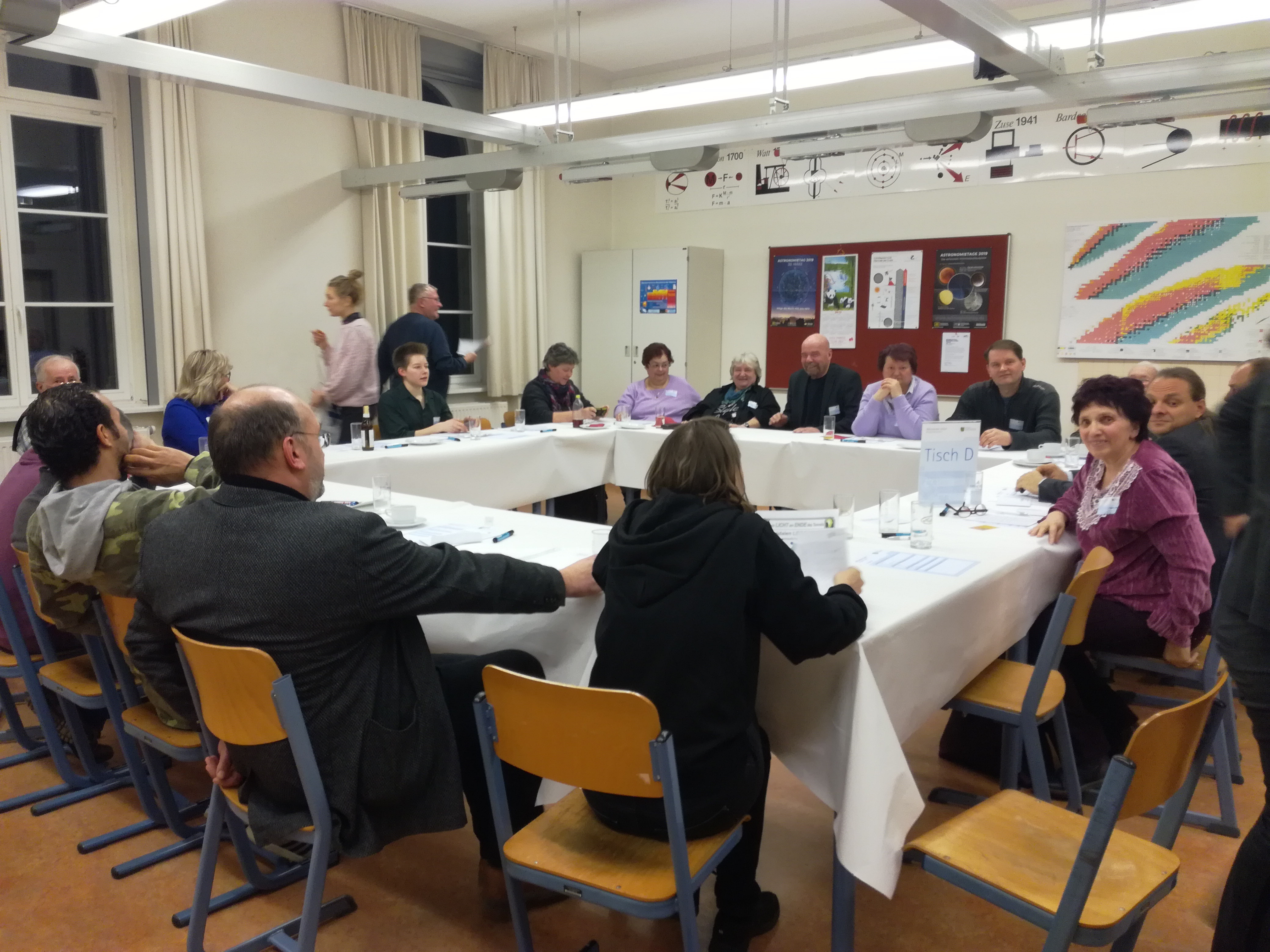 KI generiert: Das Bild zeigt eine Gruppe von Menschen, die an einem rechteckigen Tisch in einem Raum sitzen und offenbar an einem Treffen oder einer Diskussion teilnehmen. Im Hintergrund sind Poster und eine Tafel an den Wänden zu sehen.