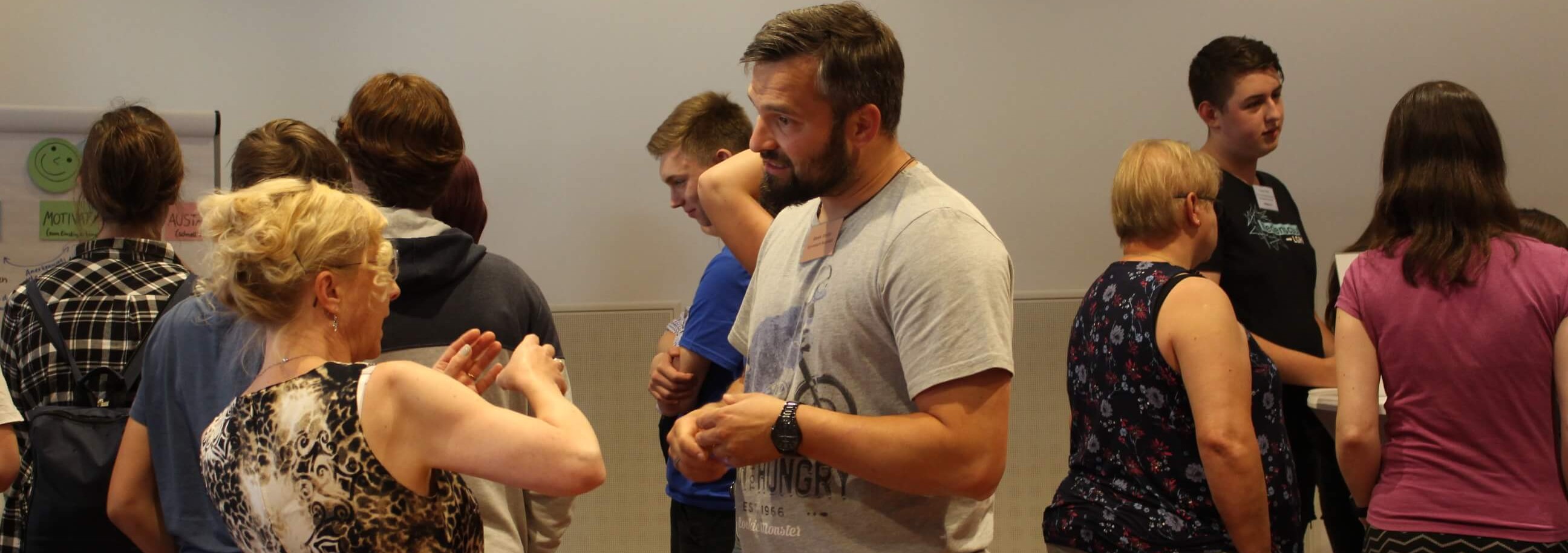 KI generiert: Das Bild zeigt eine Gruppe von Menschen, die in einem Raum in angeregter Unterhaltung sind. Einige Personen scheinen an einer Aktivität oder Diskussion beteiligt zu sein.