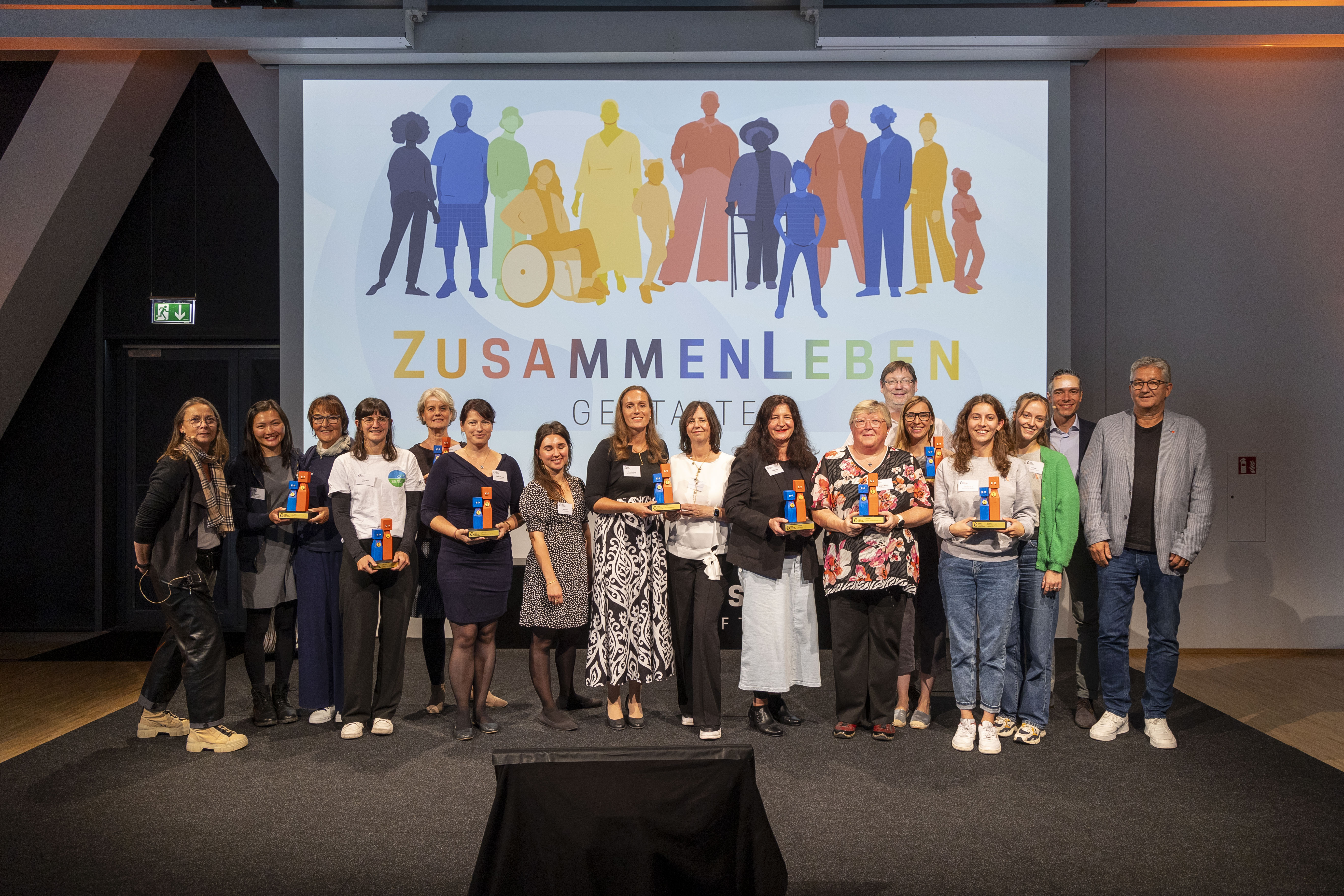 KI generiert: Eine Gruppe von Menschen steht auf einer Bühne und präsentiert Auszeichnungen oder Preise. Im Hintergrund ist eine Leinwand mit der Aufschrift "ZUSAMMENLEBEN GESTALTEN" und bunten Silhouetten zu sehen.
