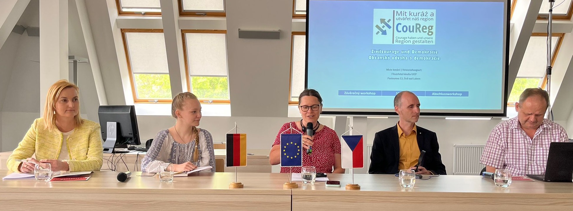 KI generiert: Fünf Personen sitzen an einem Tisch während einer Präsentation oder Diskussion. Im Hintergrund ist eine Leinwand mit dem Titel einer Veranstaltung oder eines Projekts zu sehen, und auf dem Tisch sind Flaggen von Deutschland, der EU und der Tschechischen Republik aufgestellt.