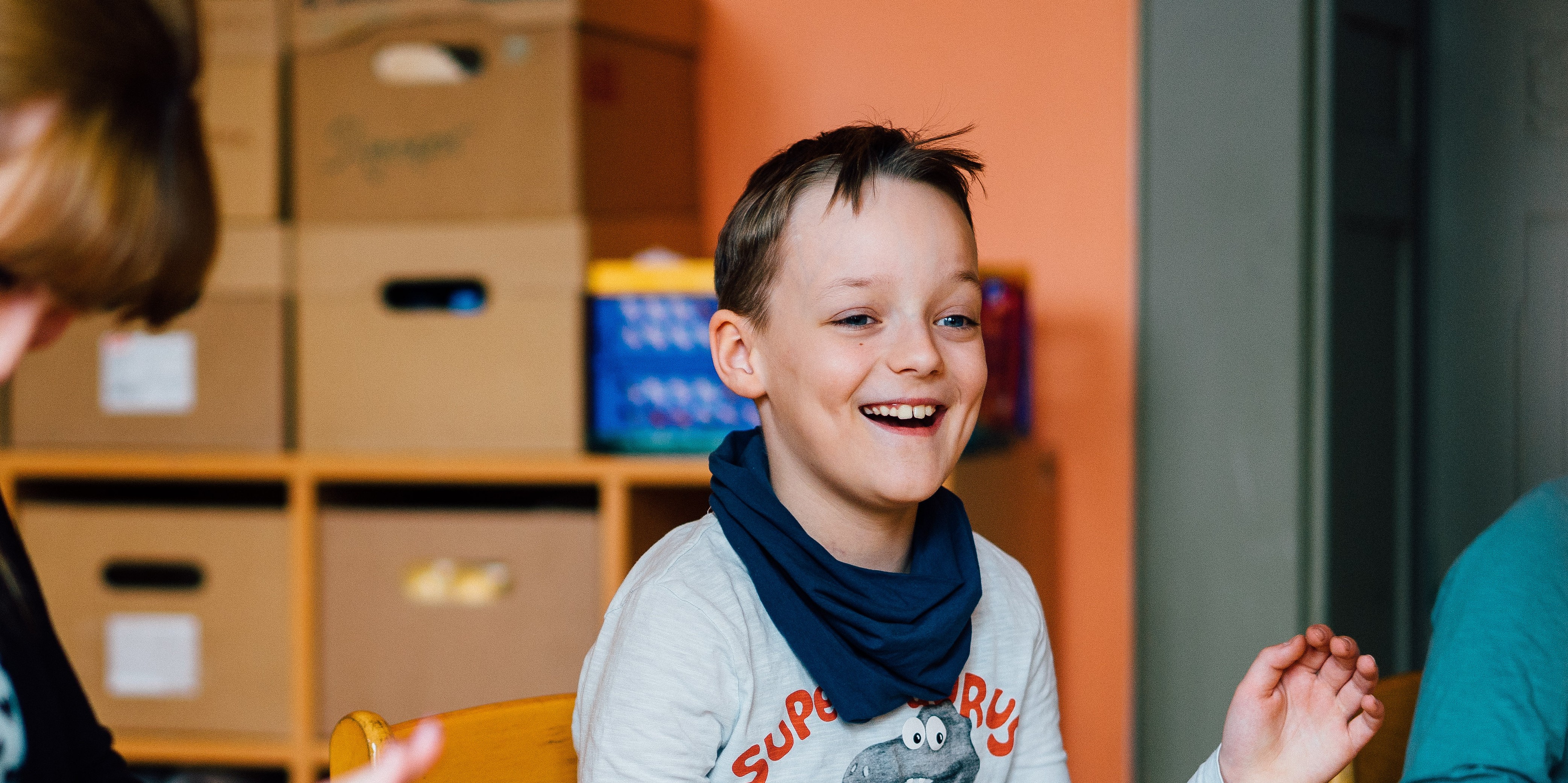 KI generiert: Ein Kind lacht fröhlich, während es an einem Tisch sitzt. Im Hintergrund sind Kisten auf Regalen zu sehen.