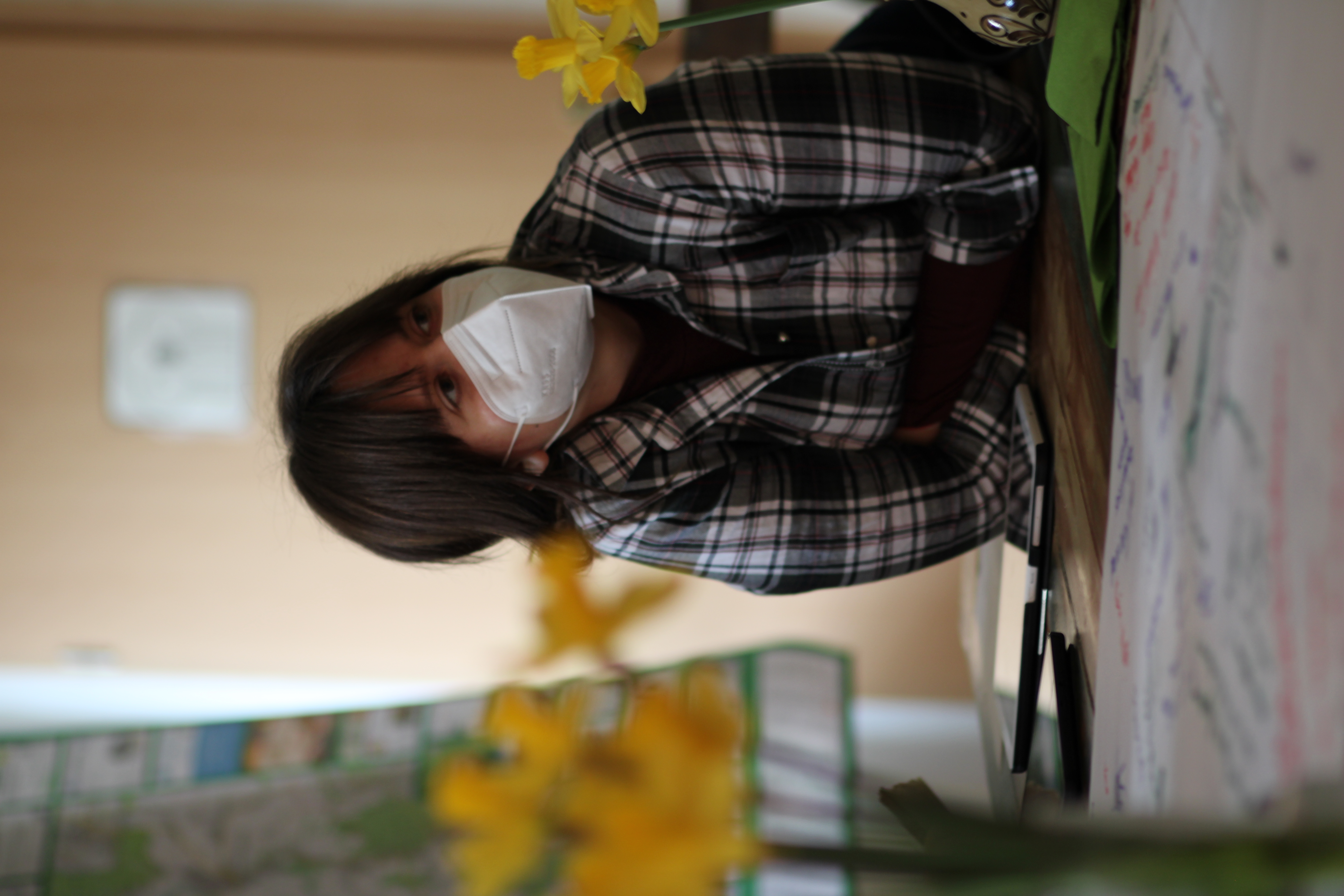 KI generiert: Eine Person mit einer Gesichtsmaske lehnt sich auf einen Tisch mit Diagrammen oder Zeichnungen, während im Vordergrund gelbe Blumen zu sehen sind. Der Hintergrund zeigt unscharf ein großes Plakat oder eine Karte.
