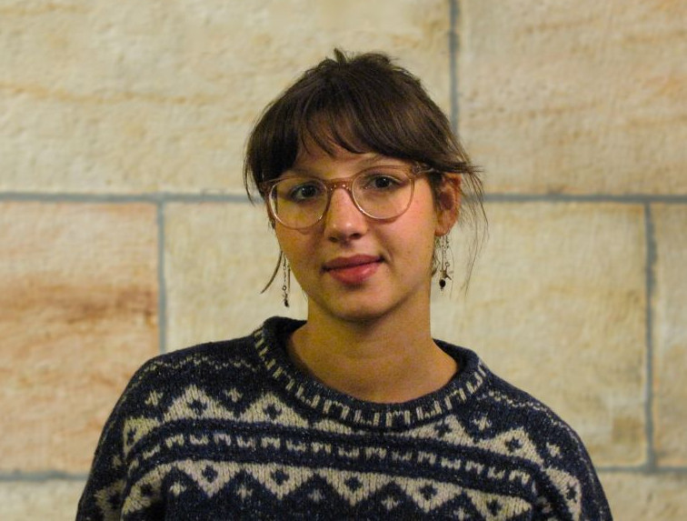 KI generiert: Das Bild zeigt eine Person mit Brille und einem gemusterten Pullover vor einem Hintergrund aus beigefarbenen Steinwänden. Die Person sieht direkt in die Kamera.