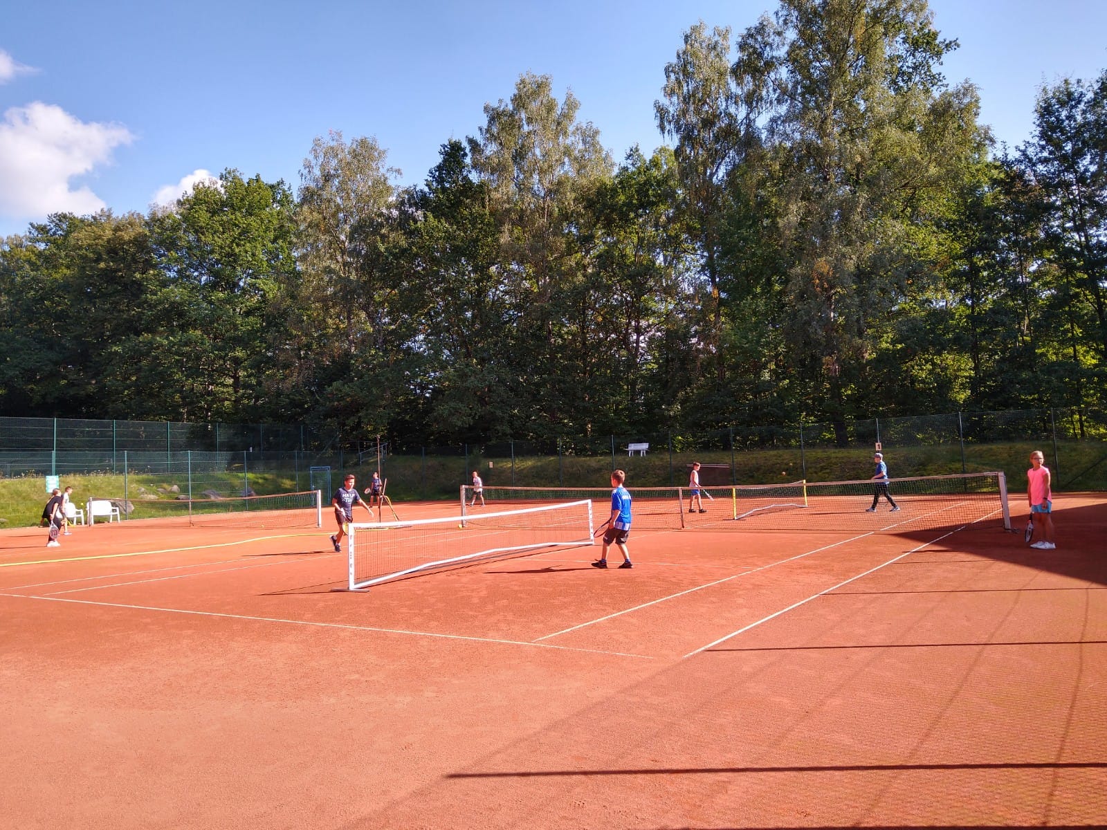 KI generiert: Das Bild zeigt einen Tennisplatz im Freien, auf dem mehrere Kinder Tennis spielen. Der Platz ist von Bäumen umgeben und es herrscht sonniges Wetter.