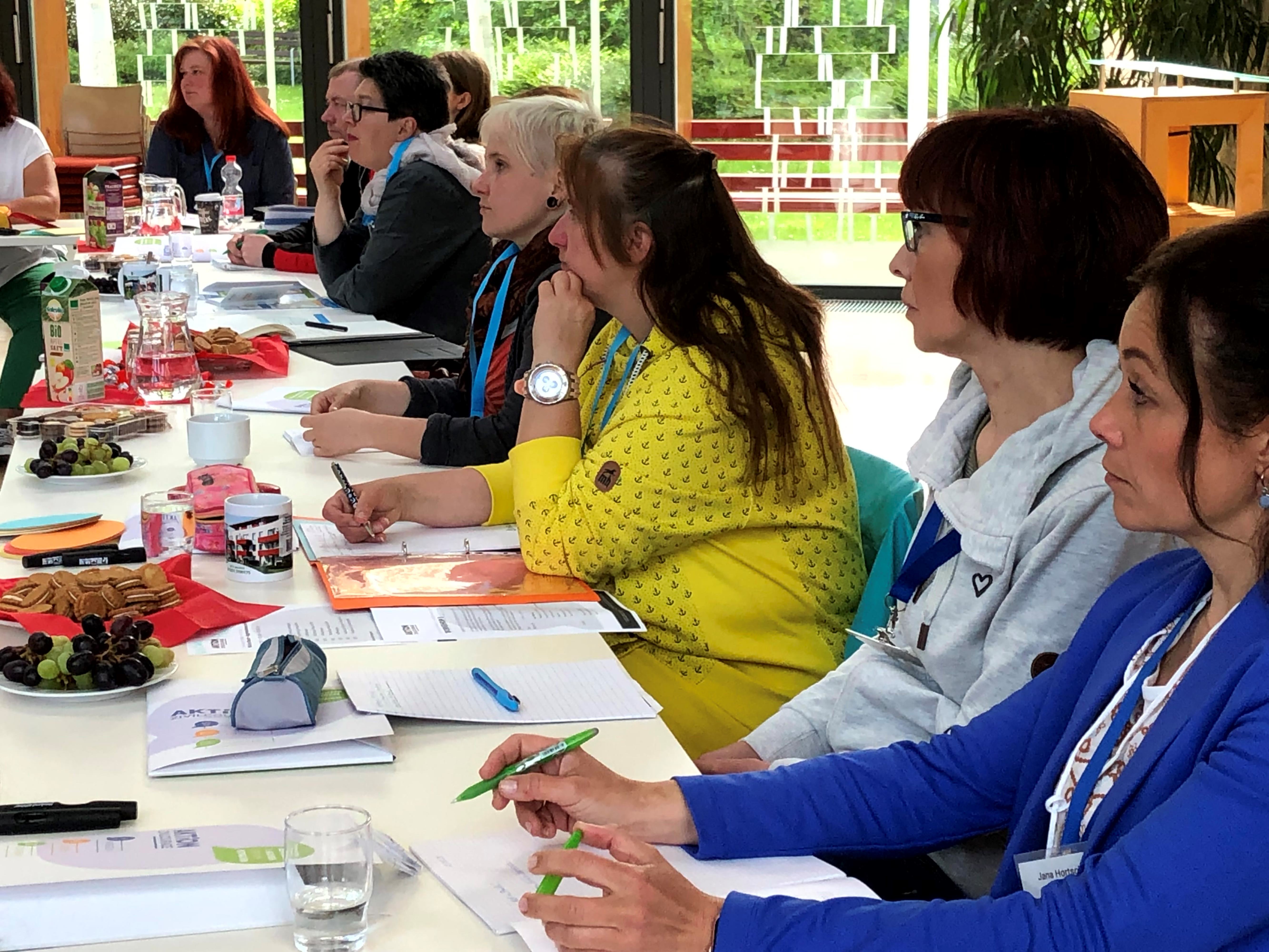 KI generiert: Eine Gruppe von Menschen sitzt an einem Tisch in einem Besprechungsraum und hört aufmerksam zu. Auf dem Tisch liegen Notizblöcke und Getränke, und es gibt einige Snacks.