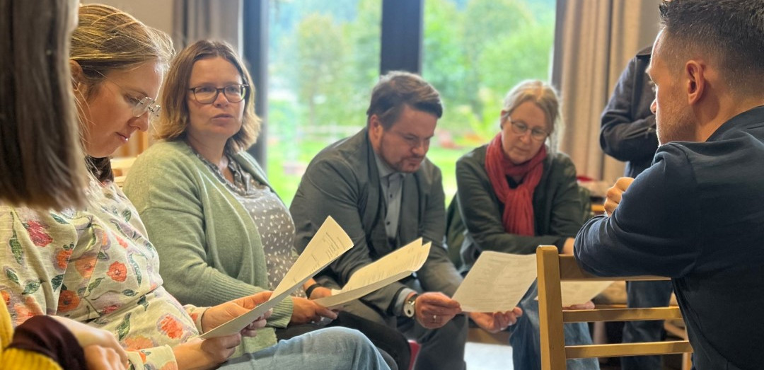 KI generiert: Eine Gruppe von Menschen sitzt zusammen und liest oder diskutiert Dokumente in einem Raum. Sie sind in eine Unterhaltung vertieft und scheinen sich in einem Meeting oder Workshop zu befinden.