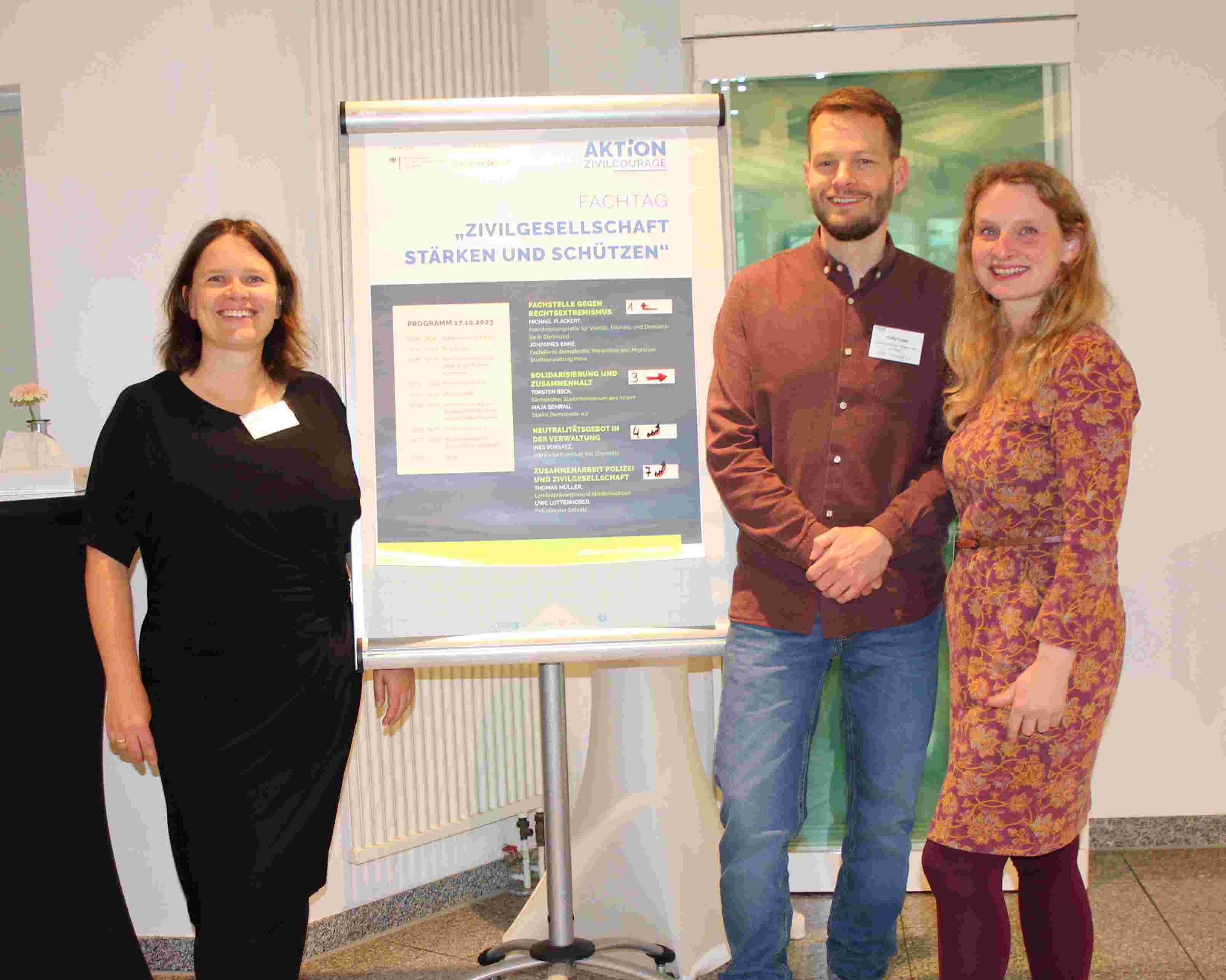 KI generiert: Das Bild zeigt drei Personen, die neben einem Plakat mit dem Titel "Fachtag Zivilgesellschaft stärken und schützen" stehen. Es handelt sich um ein professionelles Event oder eine Konferenz.