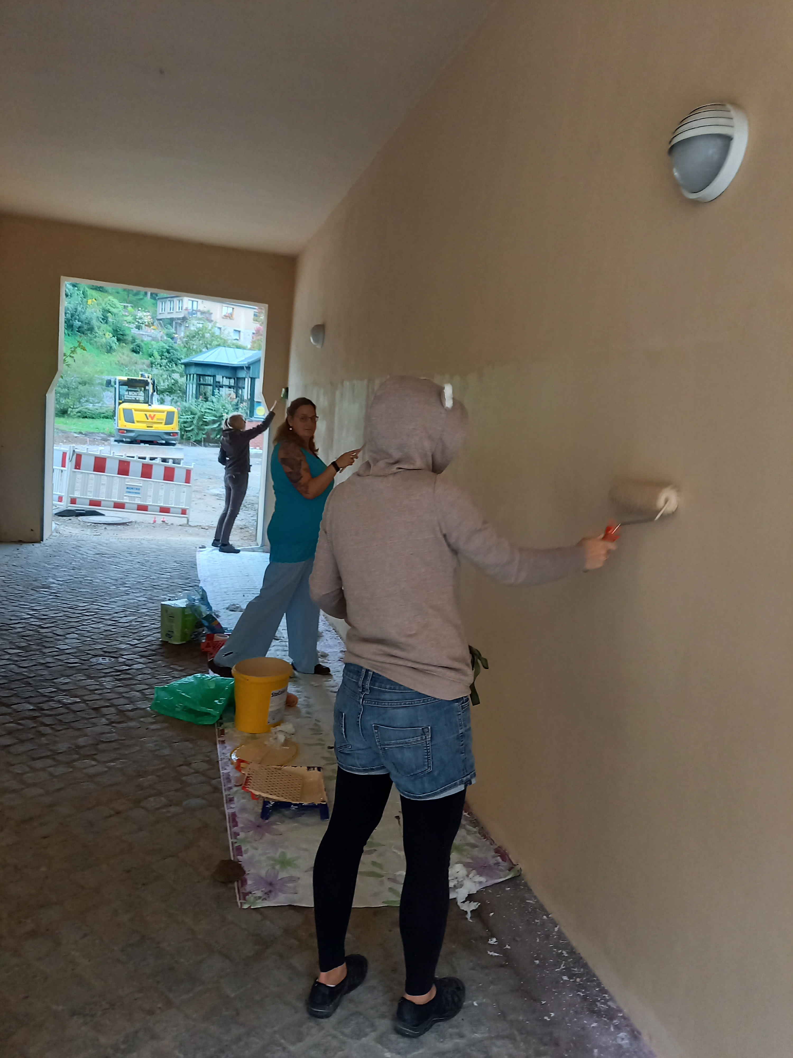 KI generiert: Das Bild zeigt mehrere Personen, die eine Wand in einem überdachten Durchgang streichen. Der Hauptinhalt ist die gemeinschaftliche Arbeit der Personen bei der Renovierung.