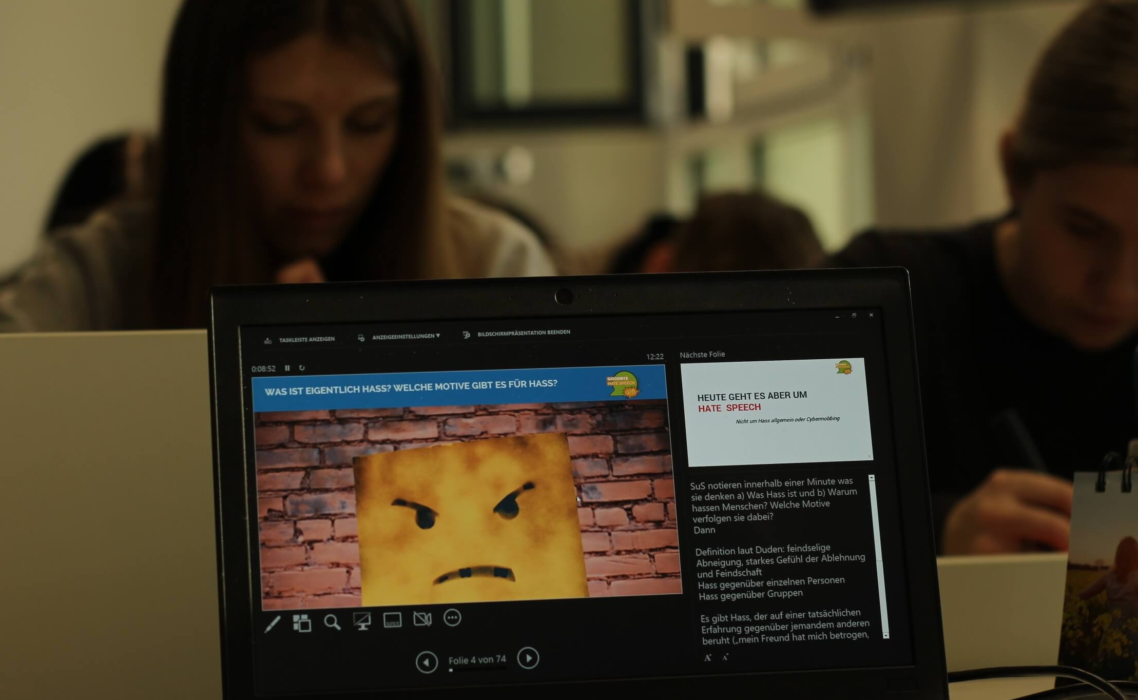 KI generiert: Das Bild zeigt einen Laptop-Bildschirm mit einer Präsentation zum Thema „Hass“, während im Hintergrund zwei Personen konzentriert arbeiten. Die Präsentation stellt Fragen zu den Motiven von Hass und scheint sich mit dem Thema Hate Speech zu beschäftigen.