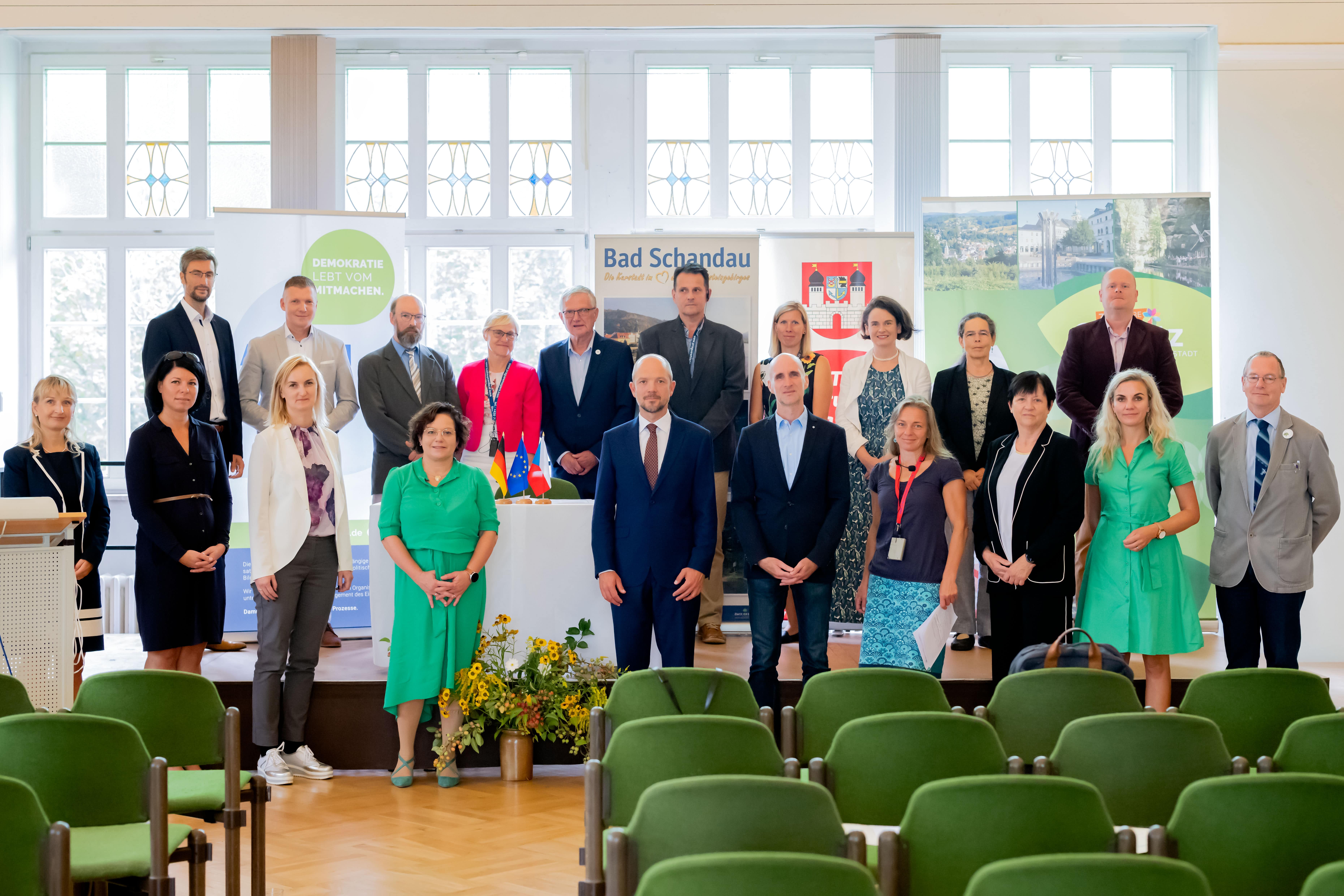 KI generiert: Eine Gruppe von Menschen steht in einer Konferenz- oder Veranstaltungsumgebung. Im Hintergrund sind Banner mit dem Text "Bad Schandau" sichtbar.