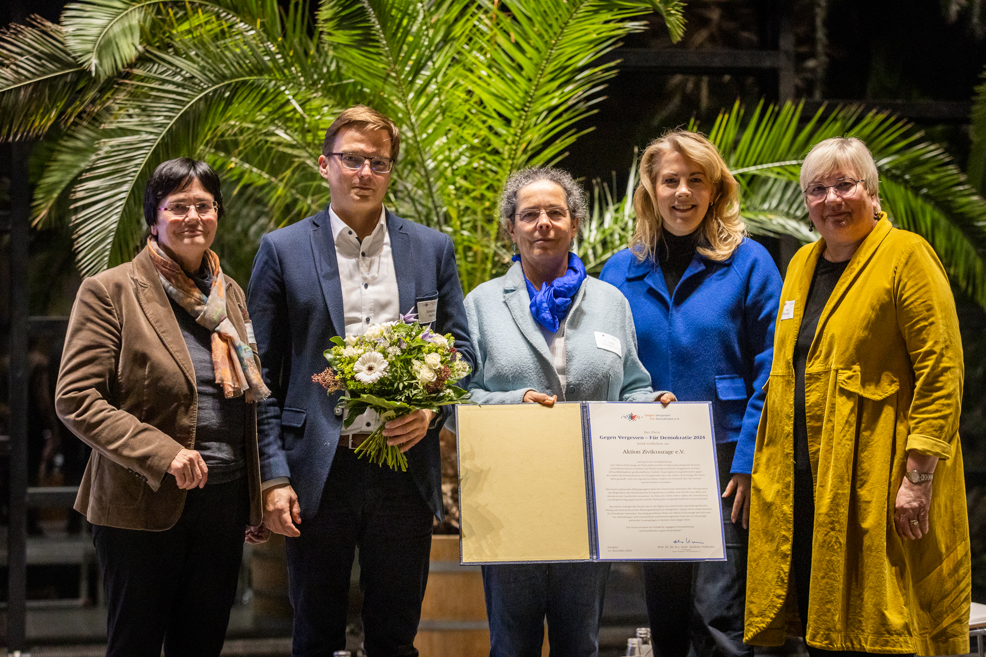 KI generiert: Eine Gruppe von fünf Personen posiert für ein Foto, wobei eine Person einen Blumenstrauß und eine andere eine Urkunde hält. Der Anlass scheint eine feierliche Auszeichnung oder Ehrung zu sein.