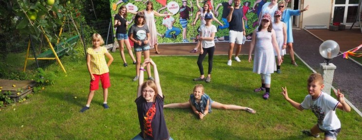 KI generiert: Das Bild zeigt eine Gruppe von Kindern und Erwachsenen, die im Freien auf einer Rasenfläche posieren. Einige Kinder führen Akrobatik oder kraftvolle Posen aus, während im Hintergrund ein buntes Plakat hängt.