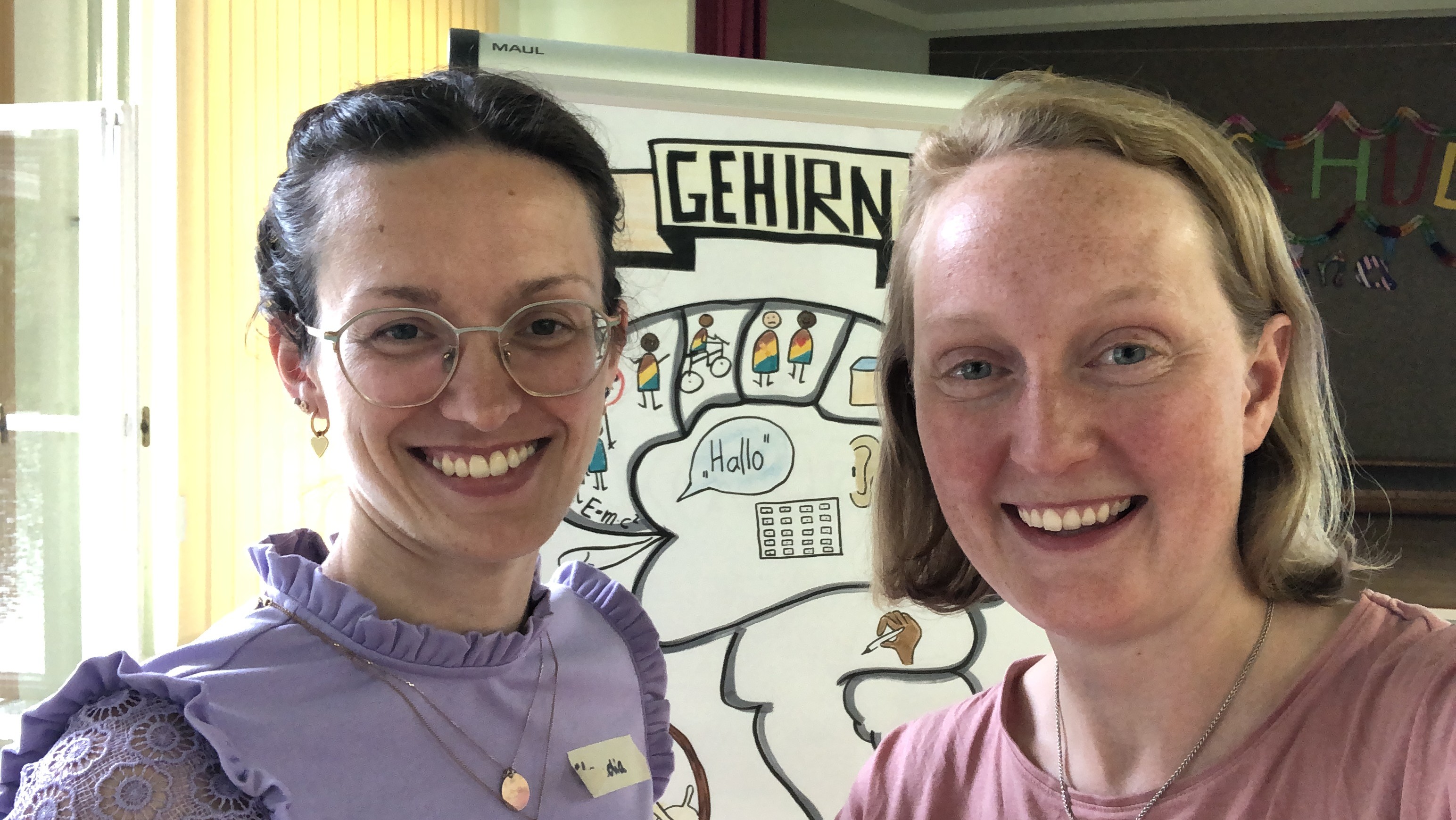 KI generiert: Auf dem Bild sind zwei lächelnde Personen zu sehen, und im Hintergrund befindet sich ein Plakat mit der Überschrift "GEHIRN", das möglicherweise Teil einer Präsentation oder eines Workshops ist. Das Umfeld deutet auf eine informelle oder lehrreiche Veranstaltung hin.