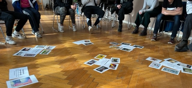 KI generiert: Eine Gruppe von Personen sitzt im Kreis auf Stühlen, während zahlreiche Papiere mit Bildern und Texten auf dem Holzboden in der Mitte verteilt sind. Der Fokus liegt auf einer Diskussion oder Aktivität, die das Material auf dem Boden einbezieht.