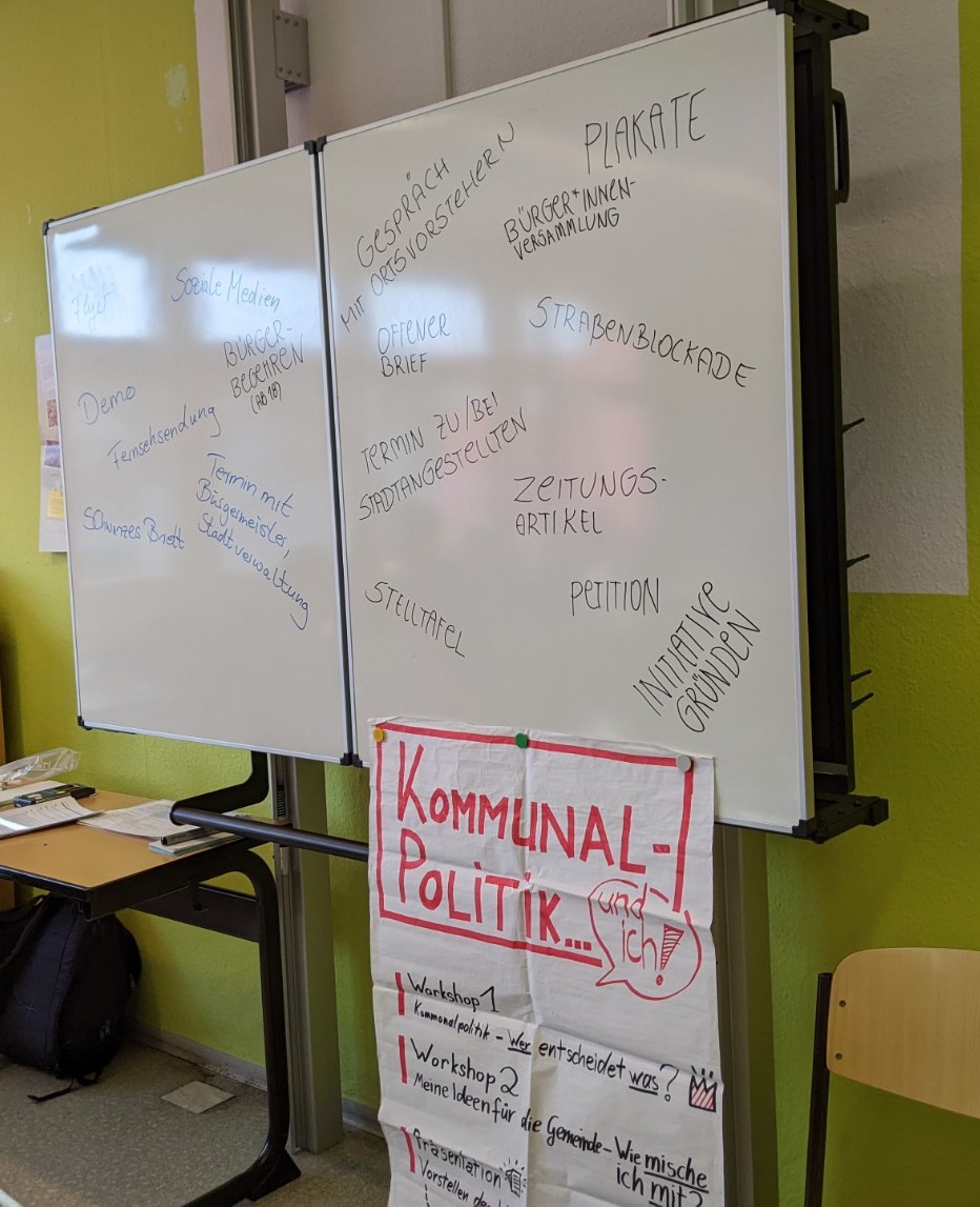 KI generiert: Das Bild zeigt eine beschriebene weiße Tafel in einem Klassenzimmer oder Seminarraum, auf der verschiedene Begriffe zur Bürgerbeteiligung stehen, wie "Straßenblockade" und "Petition". Daneben hängt ein Plakat mit dem Titel "Kommunalpolitik und ich", das Workshops zu kommunalpolitischen Themen ankündigt.