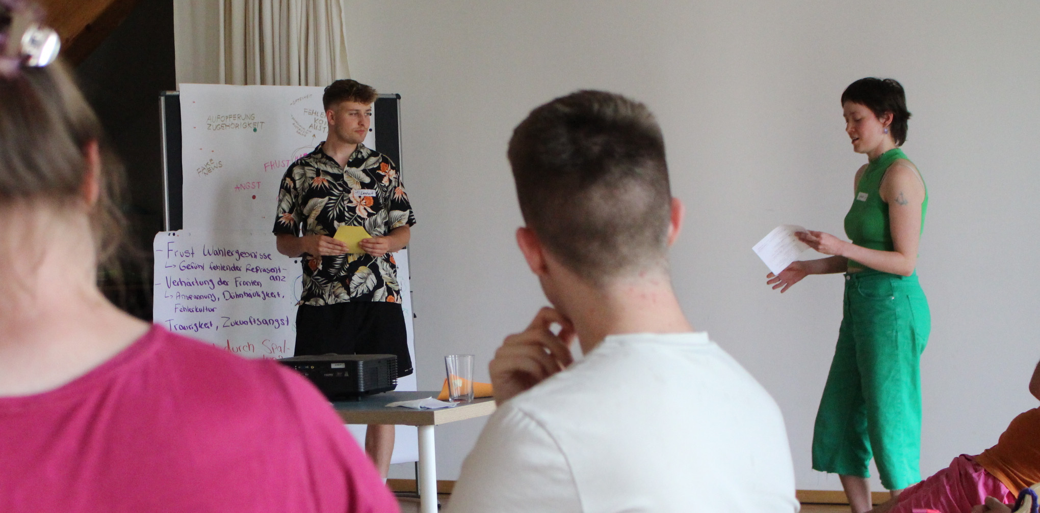 KI generiert: Das Bild zeigt zwei Personen, die vor einer kleinen Gruppe von Menschen präsentieren. Sie stehen neben einem Flipchart und halten Notizen in den Händen.