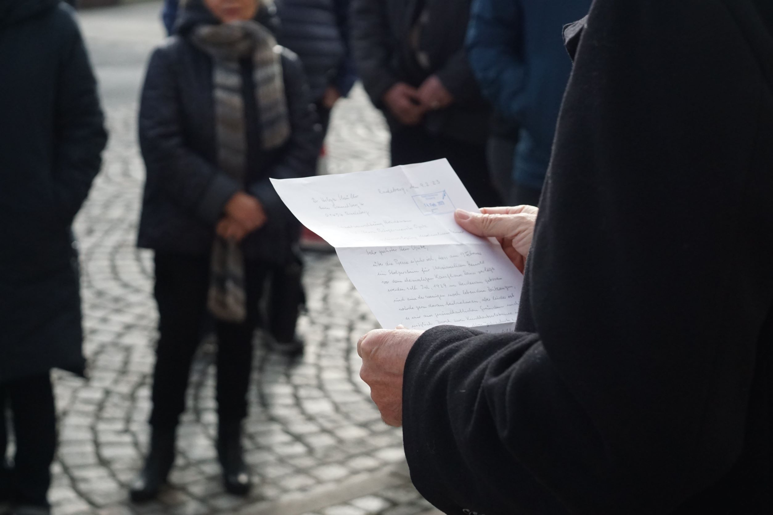 KI generiert: Eine Person hält ein handgeschriebenes Dokument in der Hand, während eine Gruppe von Menschen im Hintergrund möglicherweise einer Lesung oder Rede zuhört. Der Fokus liegt auf dem Brief, was auf eine öffentliche oder feierliche Veranstaltung hindeutet.