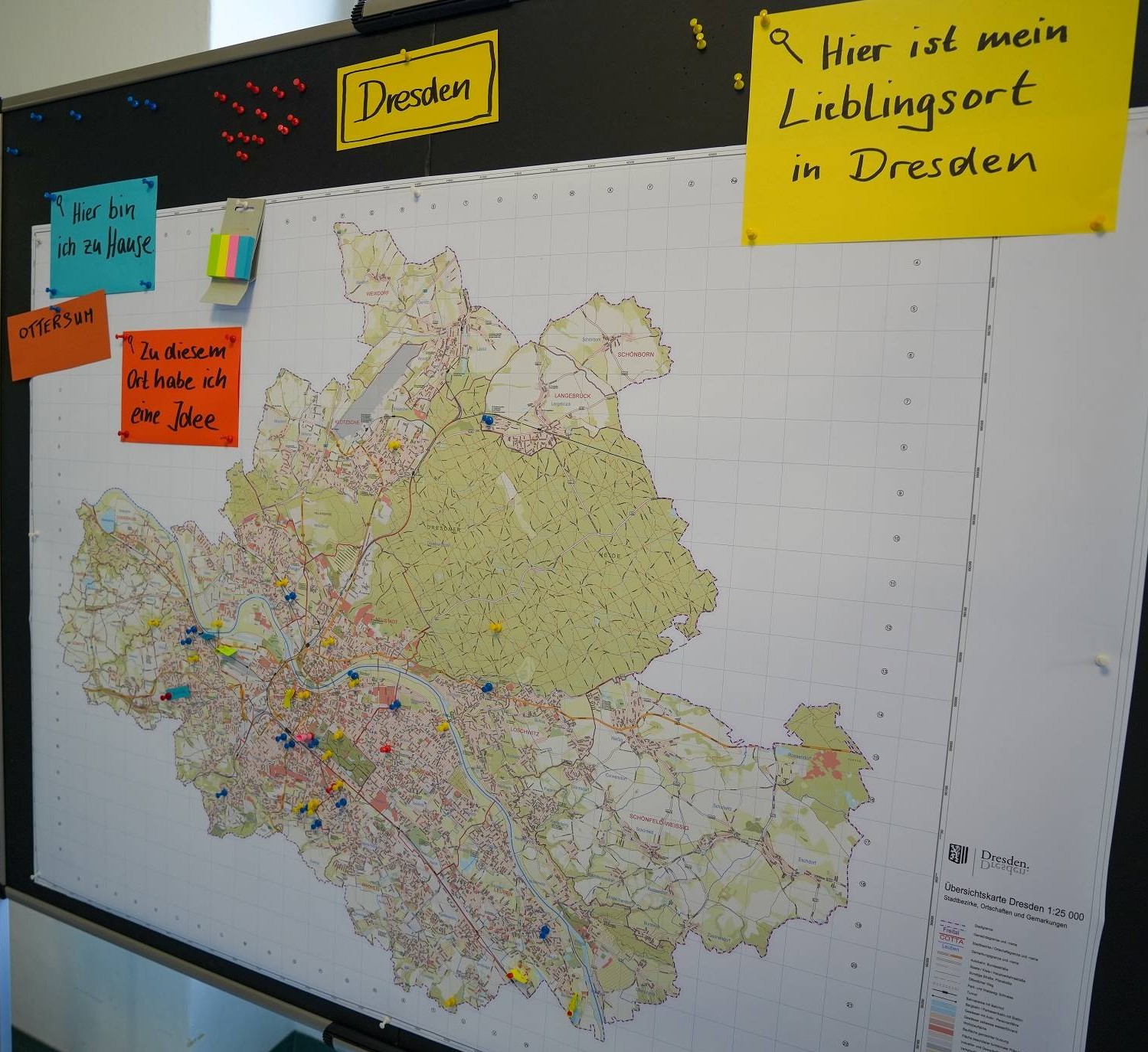 KI generiert: Das Bild zeigt eine Karte von Dresden auf einem schwarzen Brett, die mit bunten Stecknadeln markiert ist. Hinweiszettel deuten darauf hin, dass Personen Orte in Dresden als heimisch, als Lieblingsorte oder mit Ideen verbunden markiert haben.