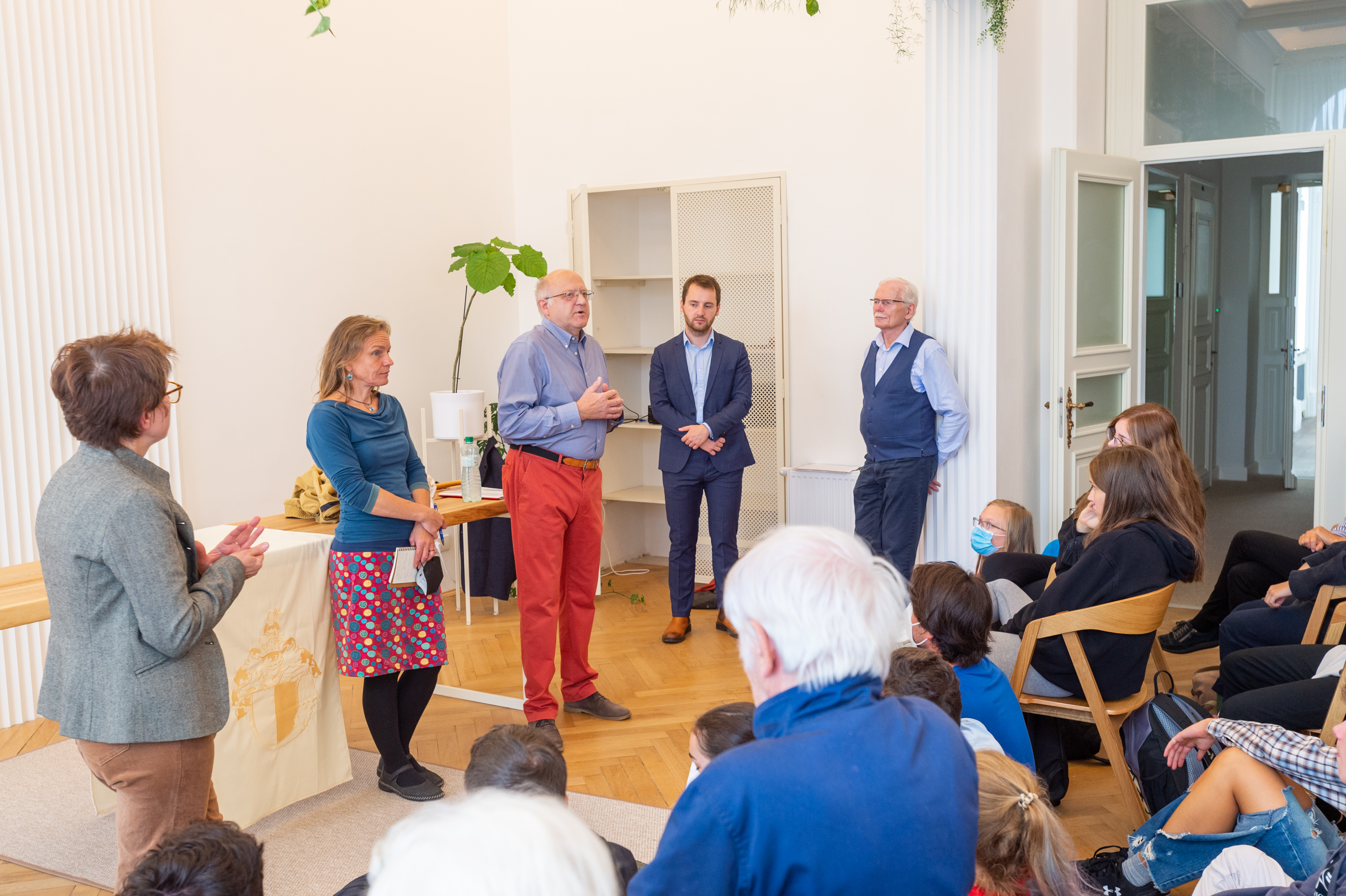 KI generiert: Das Bild zeigt eine Gruppe von Menschen bei einer Veranstaltung in einem Raum. Mehrere Erwachsene stehen im vorderen Bereich und sprechen zu einem sitzenden Publikum.