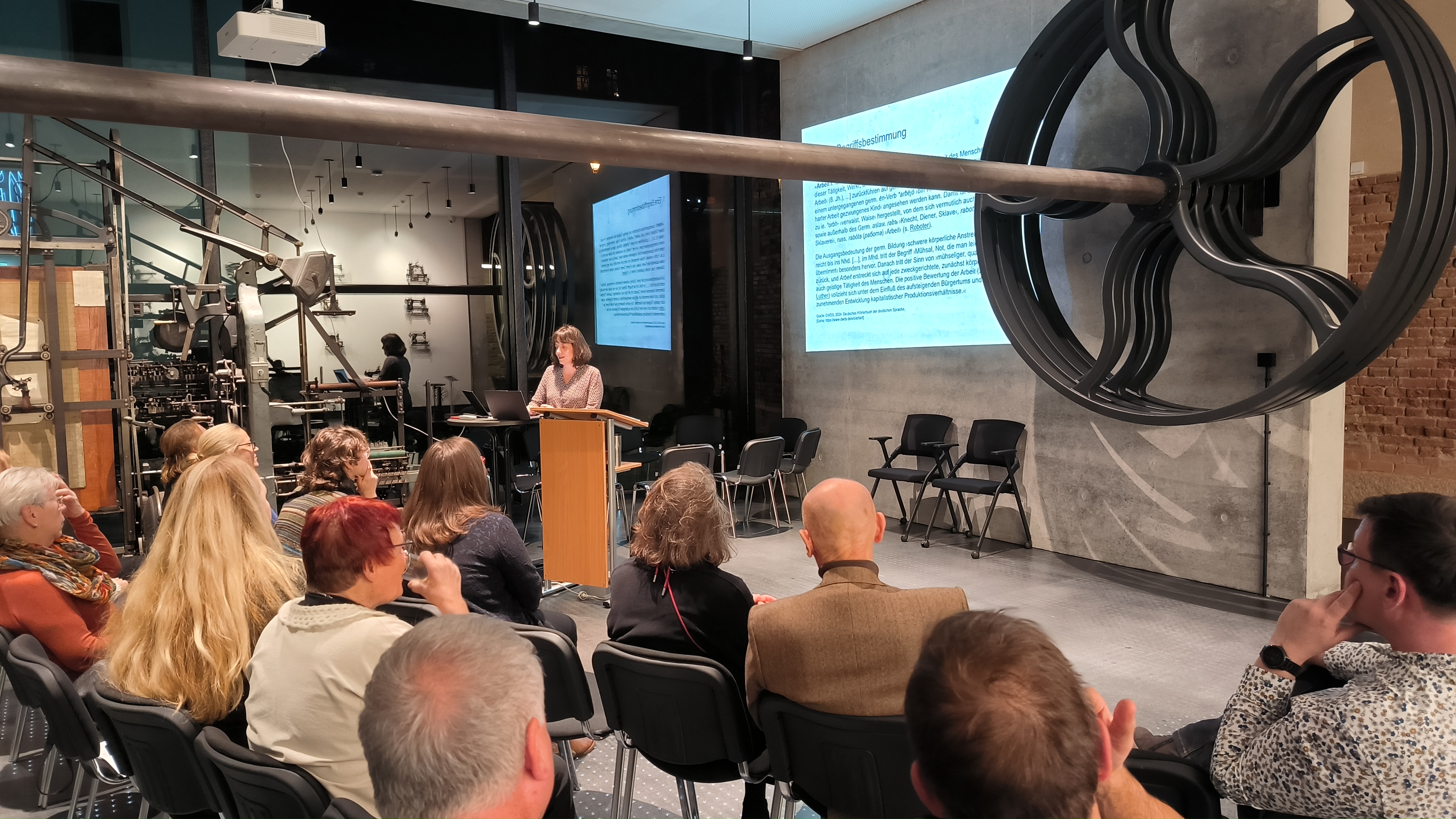 KI generiert: Das Bild zeigt eine Frau, die vor einer Gruppe von Menschen an einem Rednerpult spricht. Der Raum wirkt industriell, mit großen Maschinenteilen und einer Präsentation im Hintergrund.