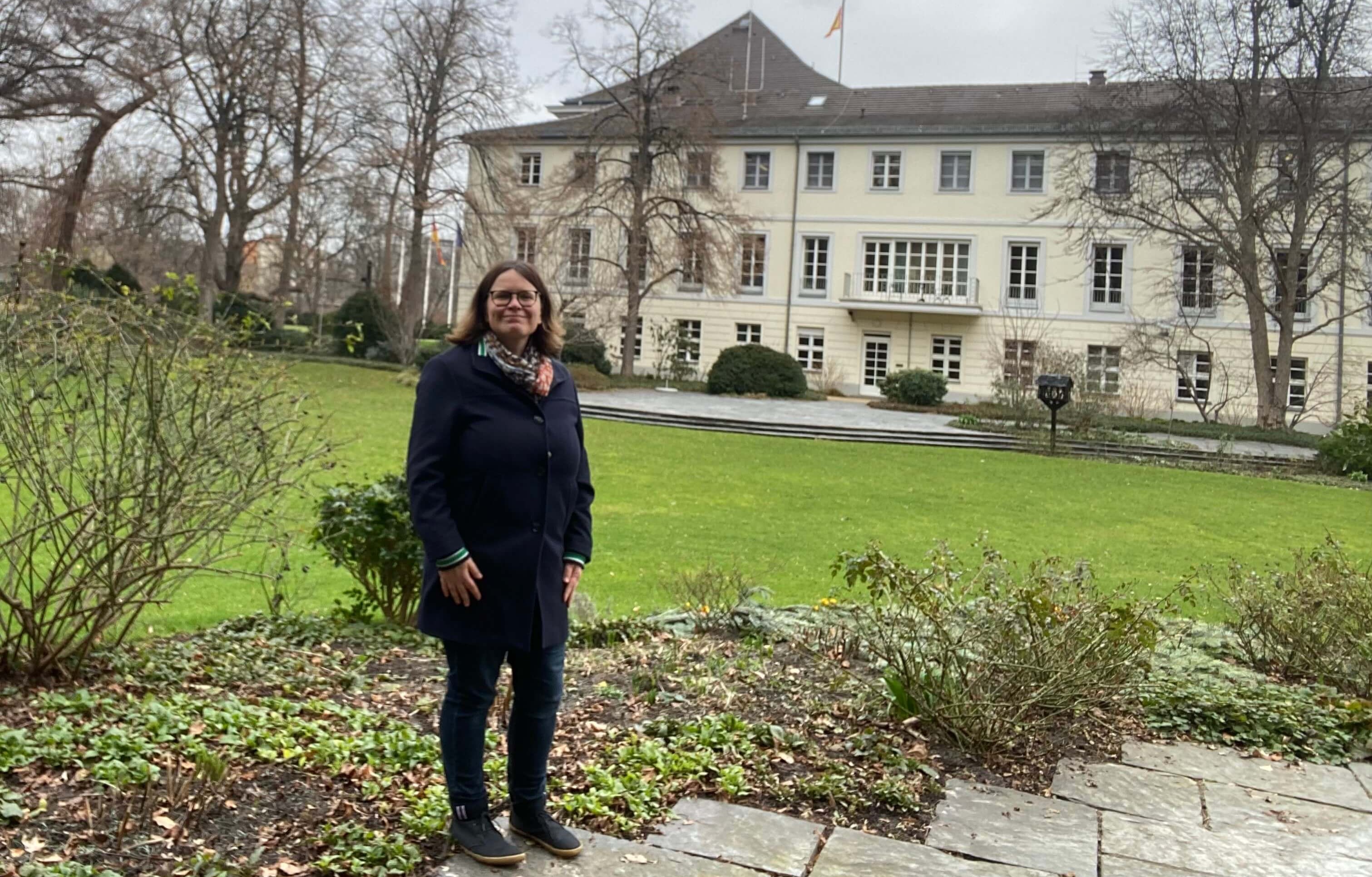 KI generiert: Eine Person steht vor einem großen, hellen Gebäude mit mehreren Fenstern und einem gepflegten Garten. Im Hintergrund ist eine Fahne auf dem Dach des Gebäudes zu sehen.