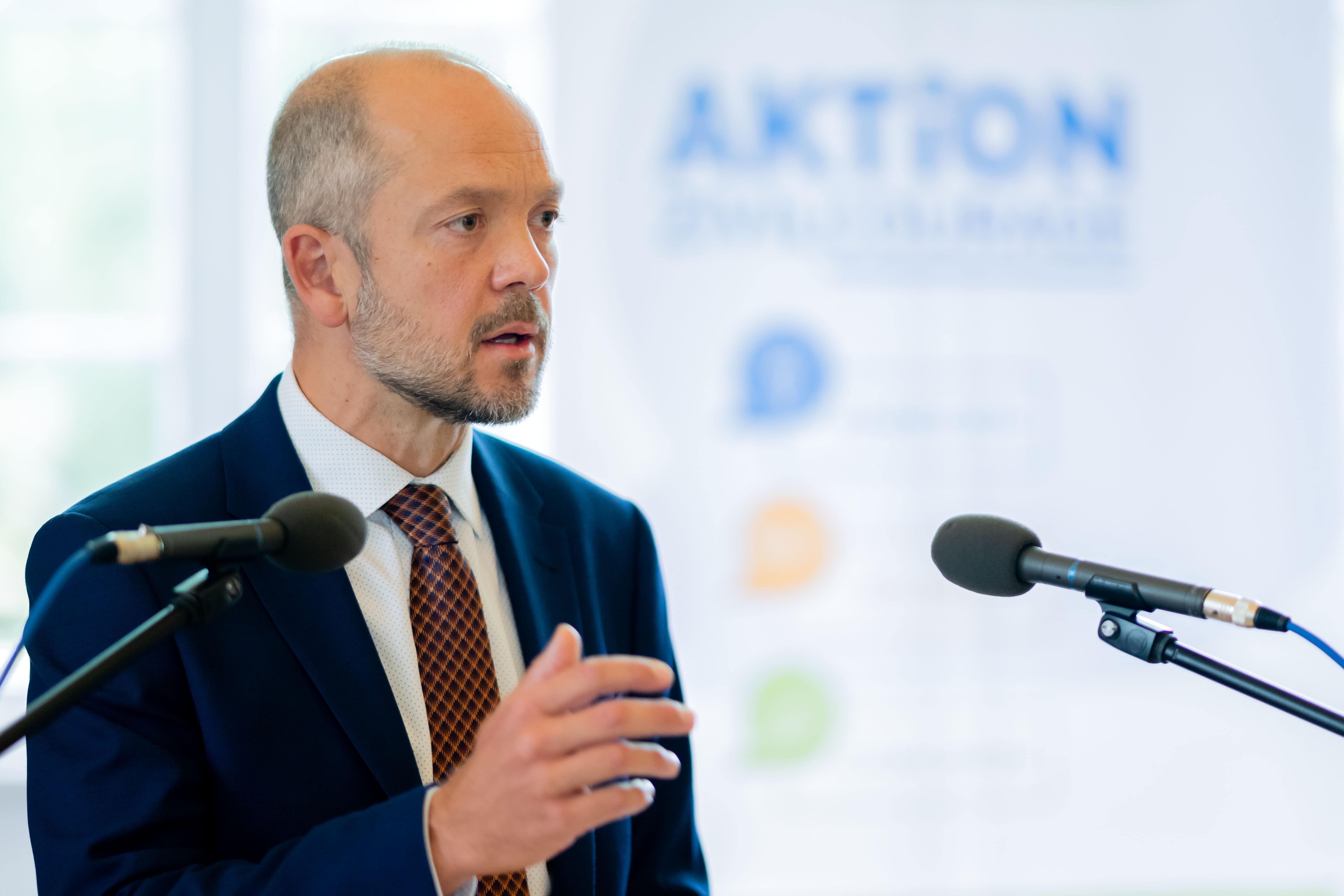 KI generiert: Ein Mann in einem Anzug hält eine Rede oder gibt ein Statement an zwei Mikrofone. Im Hintergrund ist ein Banner mit dem Wort "AKTION" zu sehen.
