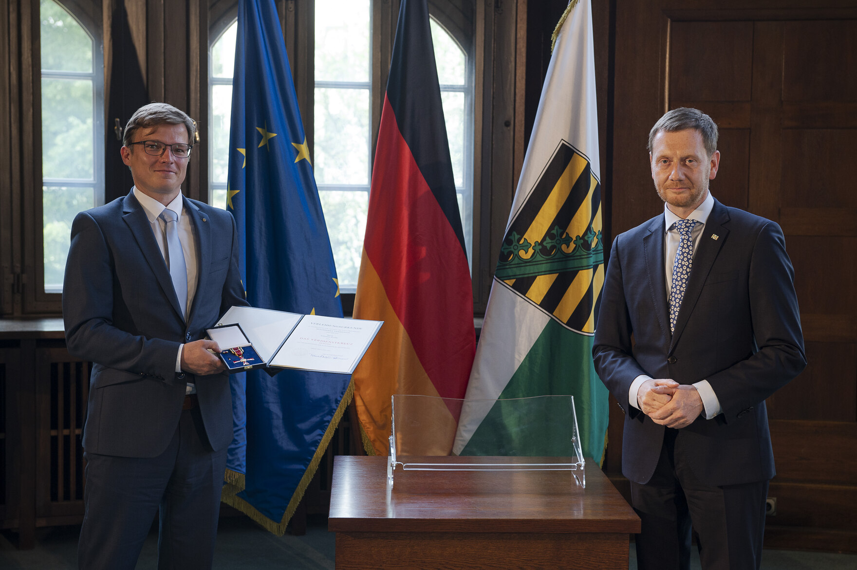 KI generiert: Zwei Männer stehen in einem formellen Raum neben einer Flaggenreihe, einer von ihnen hält eine Medaille und ein Zertifikat. Der Hauptinhalt des Bildes scheint eine offizielle Auszeichnung oder Ehrung zu sein.