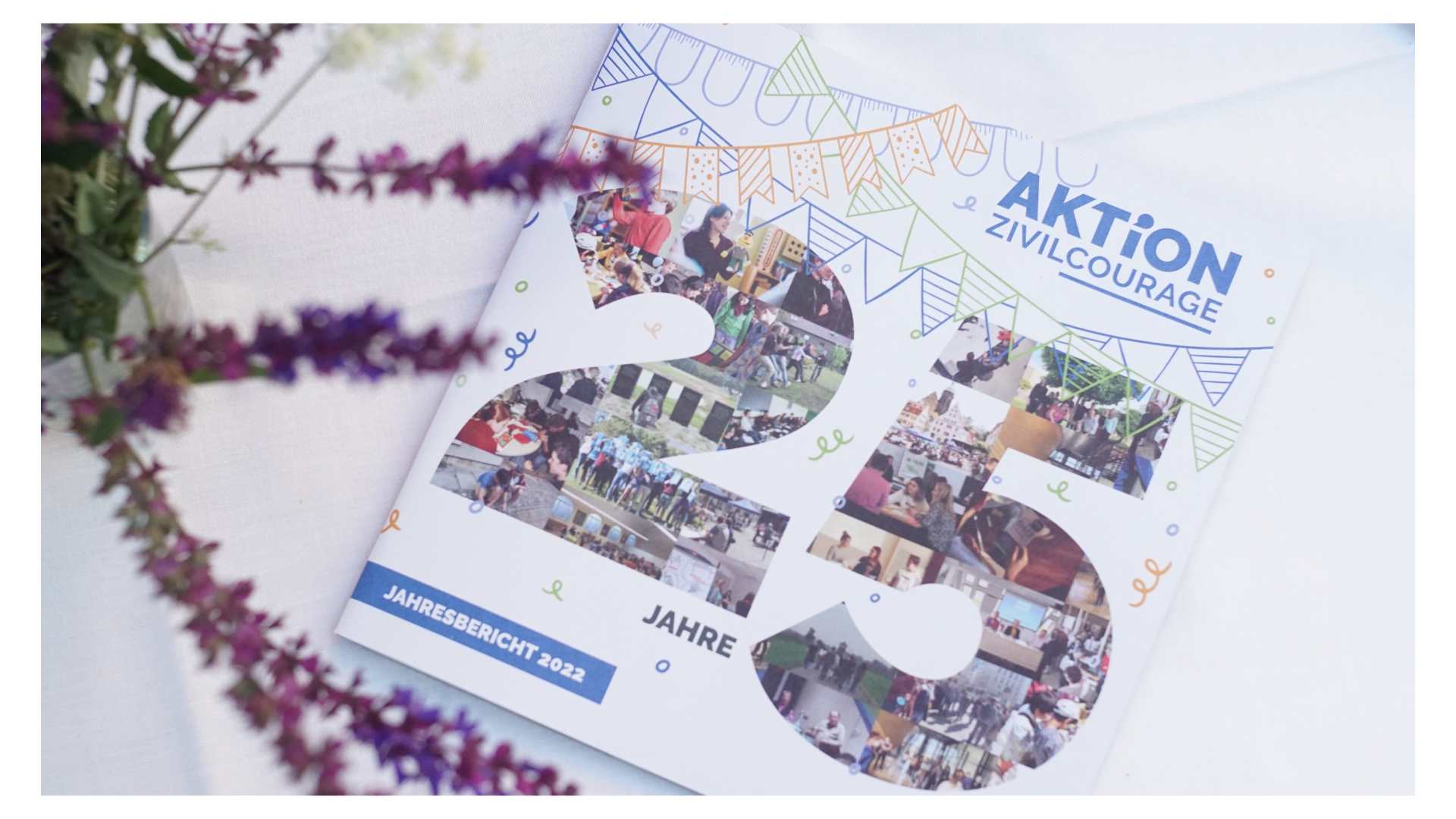 KI generiert: Das Bild zeigt den Jahresbericht 2022 der "Aktion Zivilcourage", der mit Fotos und farbigen Verzierungen den 25-jährigen Jahrestag der Organisation hervorhebt. Umgeben ist das Heft von Blumen, die eine dekorative Note hinzufügen.
