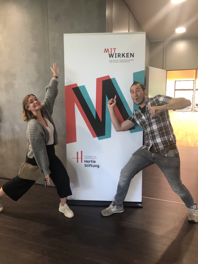 KI generiert: Zwei Personen posieren fröhlich neben einem Werbebanner der Hertie Stiftung. Der Banner trägt das Logo und den Schriftzug "MIT WIRKEN".