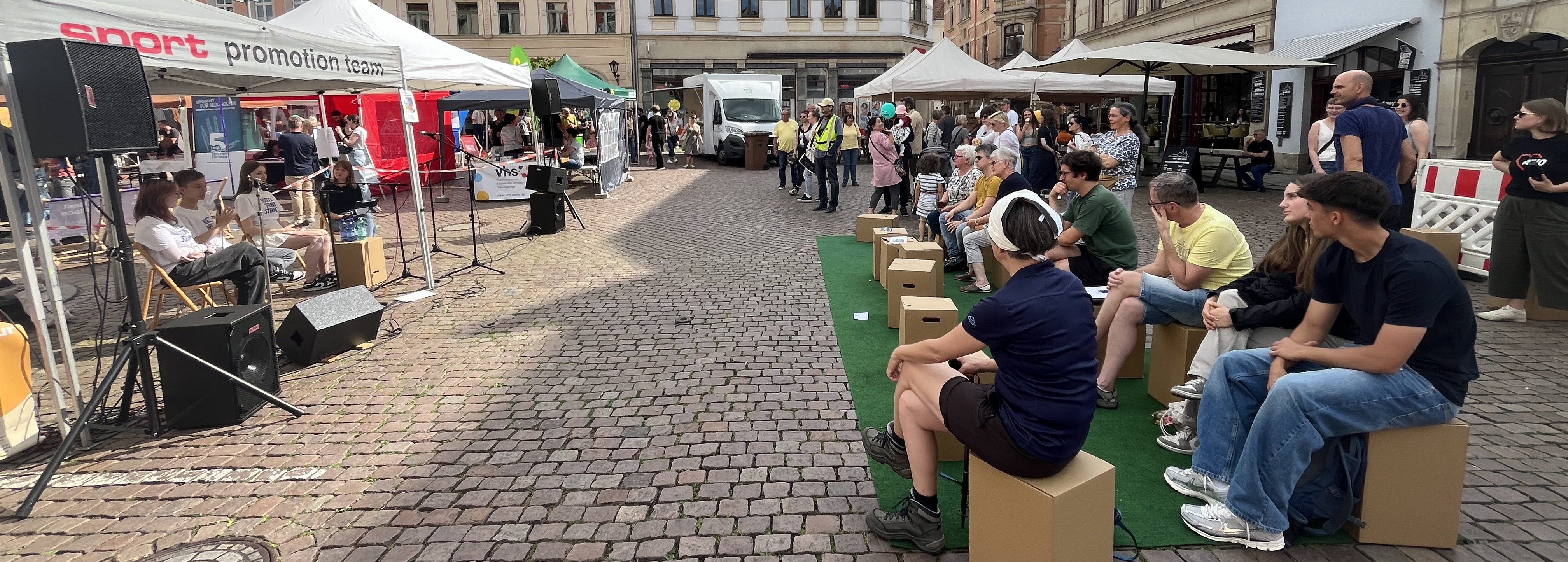 KI generiert: Das Bild zeigt eine Veranstaltung auf einem gepflasterten Platz mit mehreren Ständen und einem kleinen Publikum, das einer Präsentation oder Vorführung zuschaut. Menschen sitzen auf einfachen Sitzgelegenheiten und hören den Geschehnissen auf der Bühne zu.