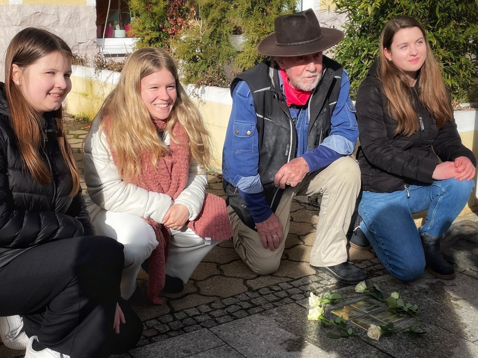 KI generiert: Das Bild zeigt vier Personen, die sich in der Nähe eines Stolpersteins auf dem Gehweg versammelt haben, umgeben von Blumen. Sie scheinen an einer Gedenkveranstaltung teilzunehmen.