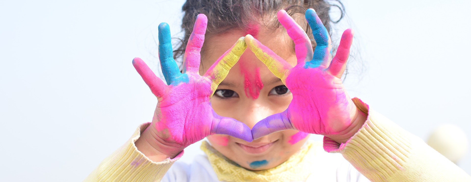 KI generiert: Ein Kind hat bunte Farben auf den Händen und im Gesicht und formt mit den Händen ein Dreieck. Im Hintergrund ist ein heller Himmel zu sehen.