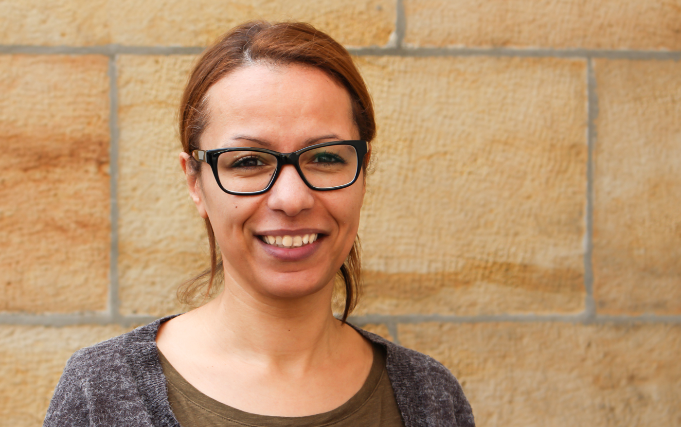 KI generiert: Das Bild zeigt eine Person, die vor einer Wand aus beigen Steinen steht und in die Kamera lächelt. Sie trägt eine Brille und wirkt freundlich.