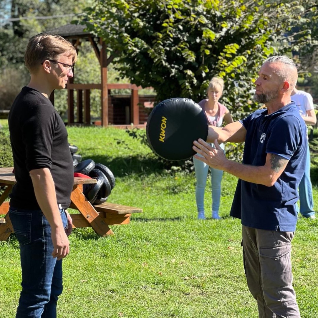 KI generiert: Das Bild zeigt zwei Männer, die sich im Freien gegenüberstehen, und einer von ihnen hält einen schwarzen Trainingsball in den Händen. Im Hintergrund sind eine Frau und ein grüner Rasen zu sehen.