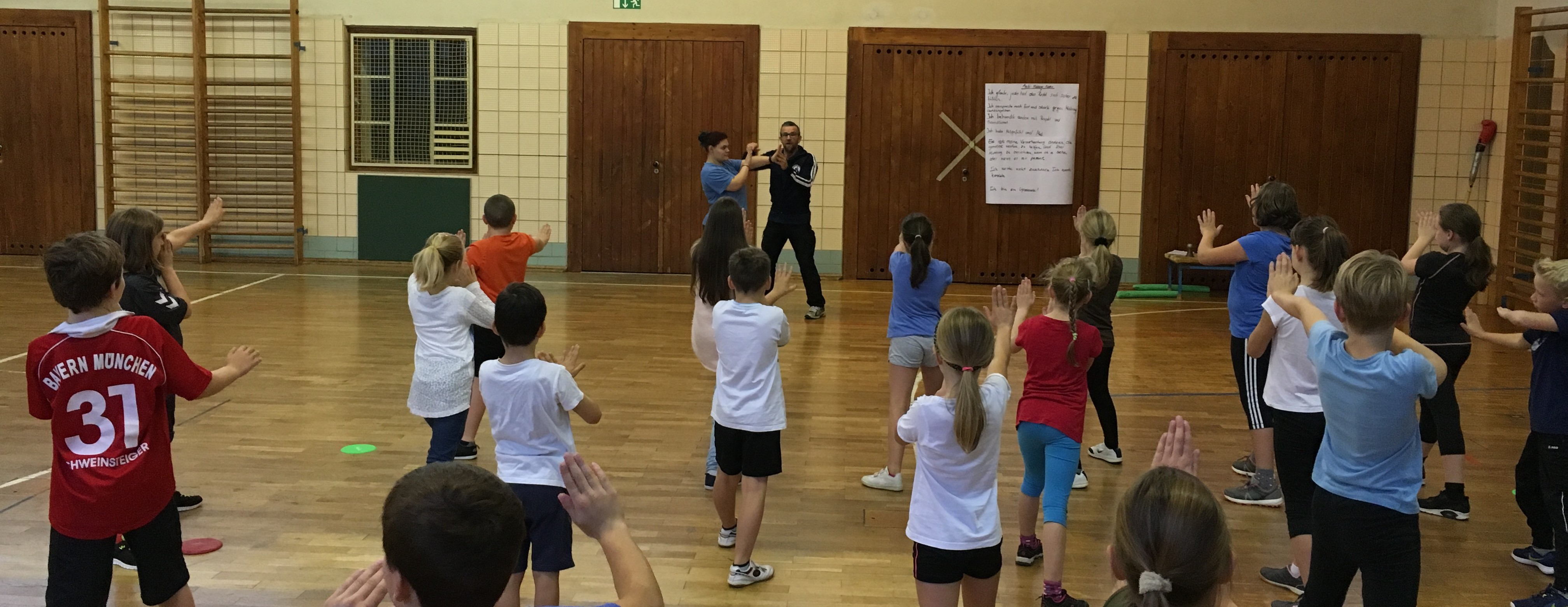 KI generiert: Das Bild zeigt eine Gruppe von Kindern in einer Turnhalle, die unter Anleitung von zwei Erwachsenen an einer sportlichen Aktivität teilnehmen. Die Kinder stehen in einer Formation und scheinen Übungen oder Bewegungen auszuführen.