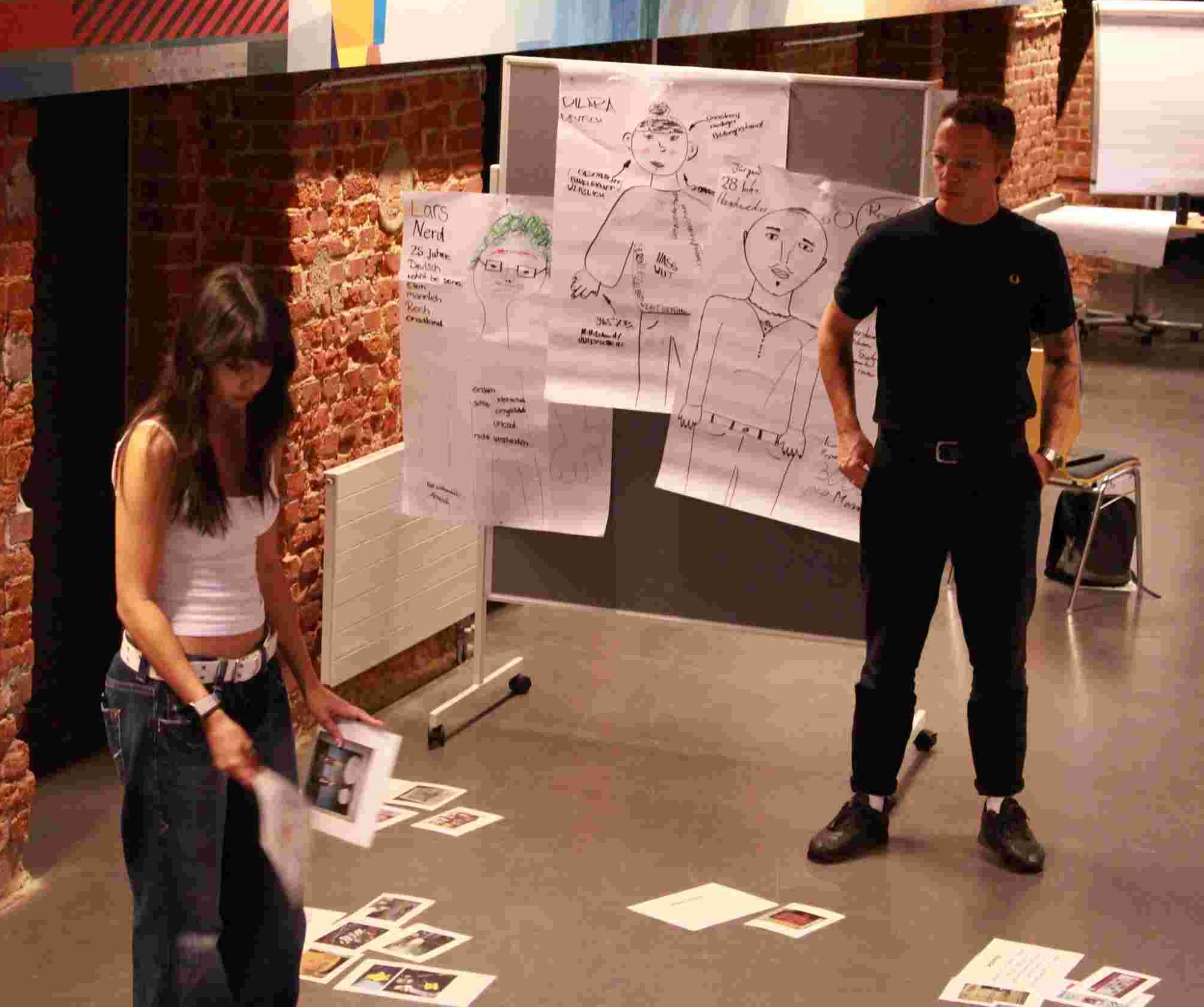 KI generiert: Das Bild zeigt zwei Personen in einem Raum mit Ziegelwänden, die scheinbar an einer Präsentation oder einem Workshop teilnehmen. Im Hintergrund sind mehrere Poster mit Zeichnungen und Notizen zu sehen, die an einer Stellwand befestigt sind.